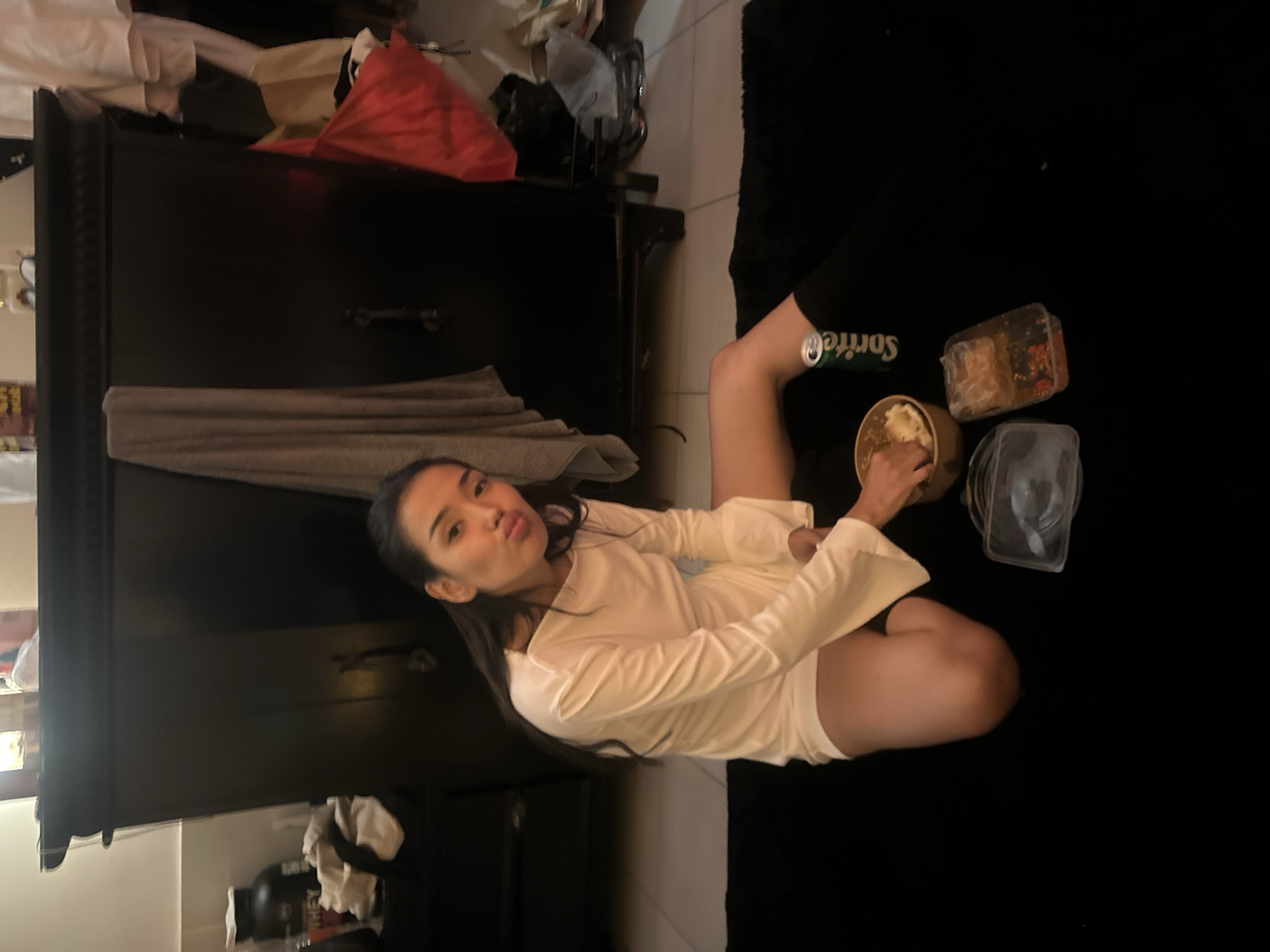 The image shows a person sitting on a black surface, possibly a bed or floor, with their legs crossed. They are wearing a long-sleeved white top and shorts, and are holding a bowl of food. Next to them are containers of food and a can of Sprite. In the background is a dark-colored wardrobe. The image appears to be taken indoors. The location, North Kuta, Indonesia, is based on available information but cannot be confirmed solely from this image.