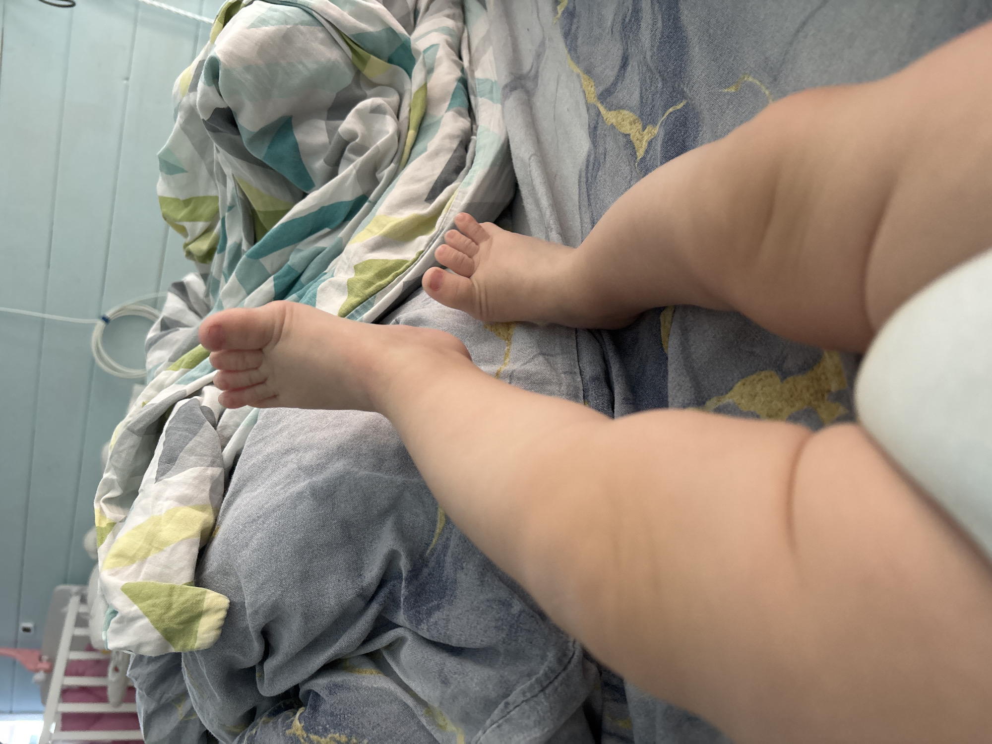 The image shows a close-up view of a baby's legs and feet lying on a bed with patterned bedding. The baby is positioned on its back, with its legs extended. The bedding is a mix of blues, greens, whites, and yellows. In the background, a crib with pink and white bars is visible, along with a light blue wooden wall. The overall impression is one of a domestic setting, likely a nursery or bedroom. The location of Раздолье, Russia, cannot be determined from the visual information in the image.