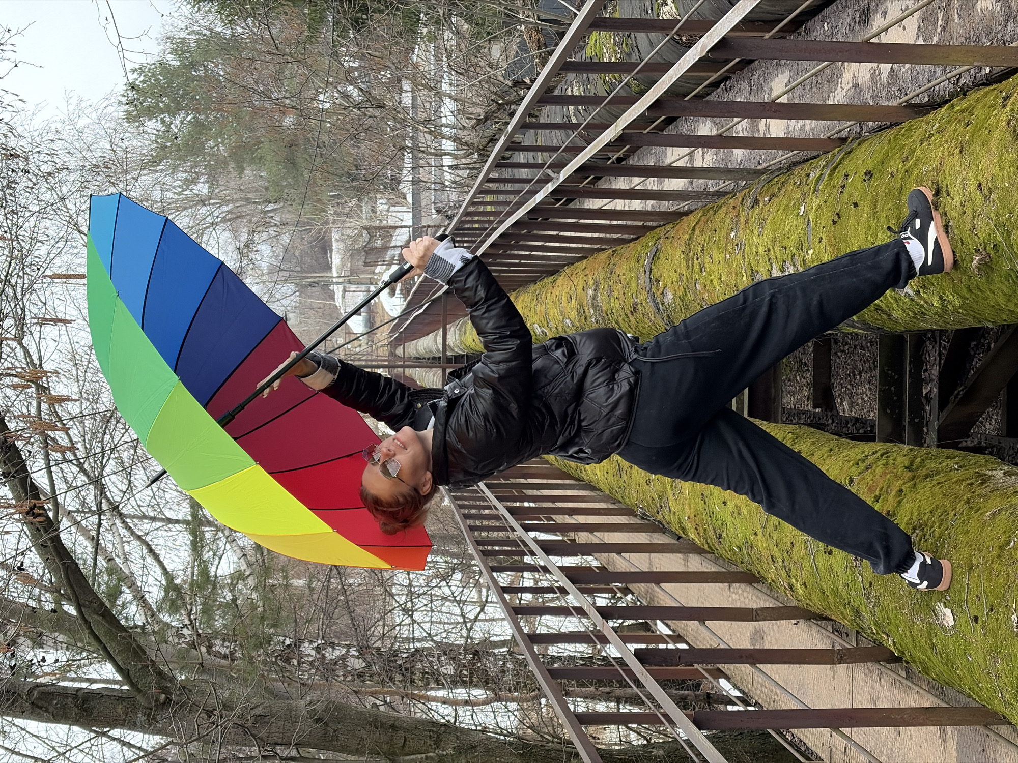 A woman in black is poised on a moss-covered pipe near a metal ladder structure in what appears to be an industrial or outdoor setting in Chaikovsky, Russia. She is holding a multi-colored umbrella. Trees and buildings are visible in the background.