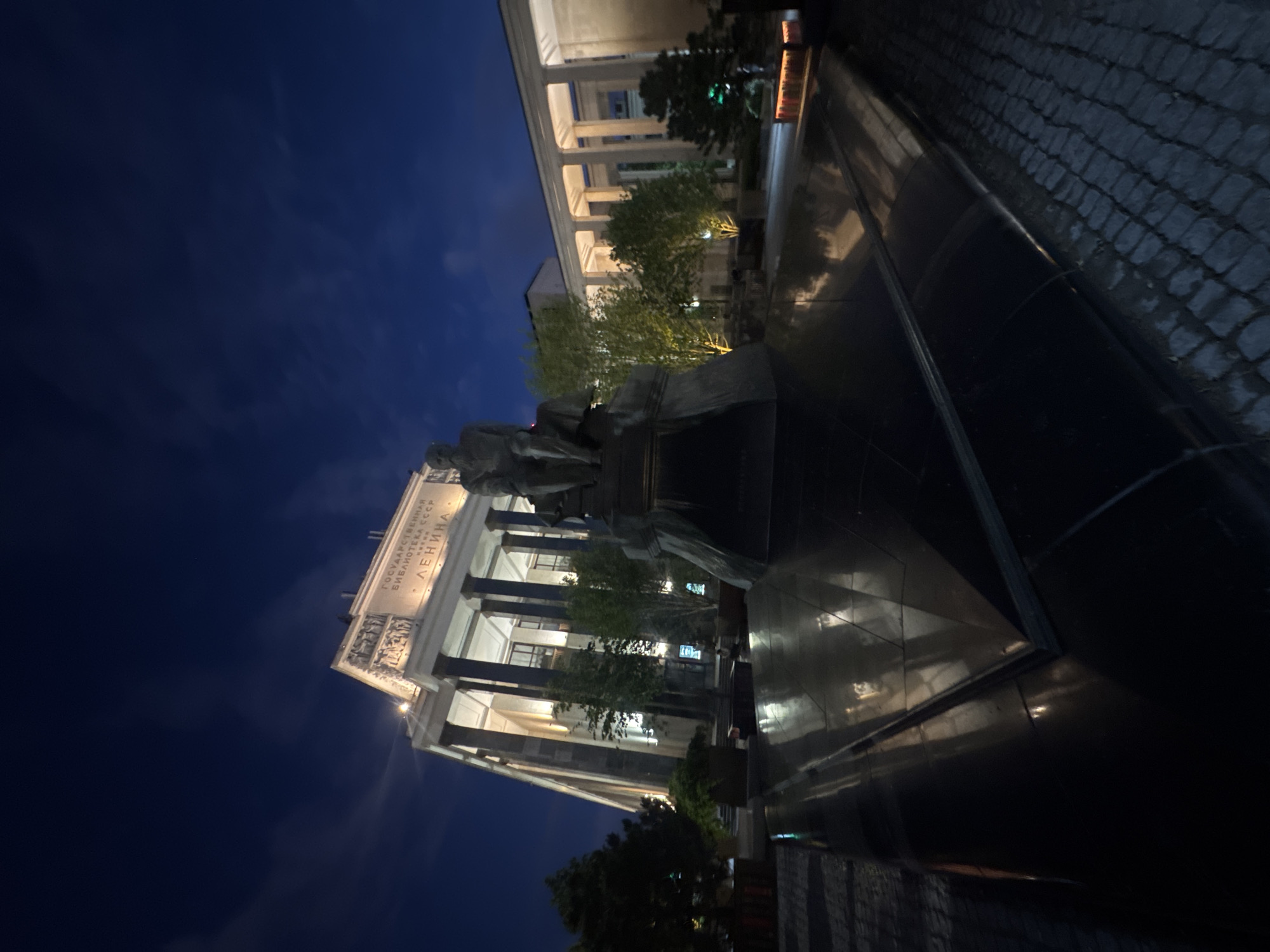 The photograph, taken at night, shows the monument to Vladimir Lenin in front of the Russian State Library in Moscow, Russia. The imposing neoclassical building, illuminated from below, features a grand facade with classical columns. The statue of Lenin is positioned on a dark, stepped pedestal in the foreground, its silhouette stark against the night sky. The ground is paved with cobblestones, and reflective surfaces of the monument's base create a mirror-like effect, reflecting the lights of the building and the sky. Lush greenery surrounds the monument and building, with trees and planters adding to the urban landscape. The sky is dark blue, with wisps of clouds illuminated by the ambient light. No people are clearly visible in the frame.