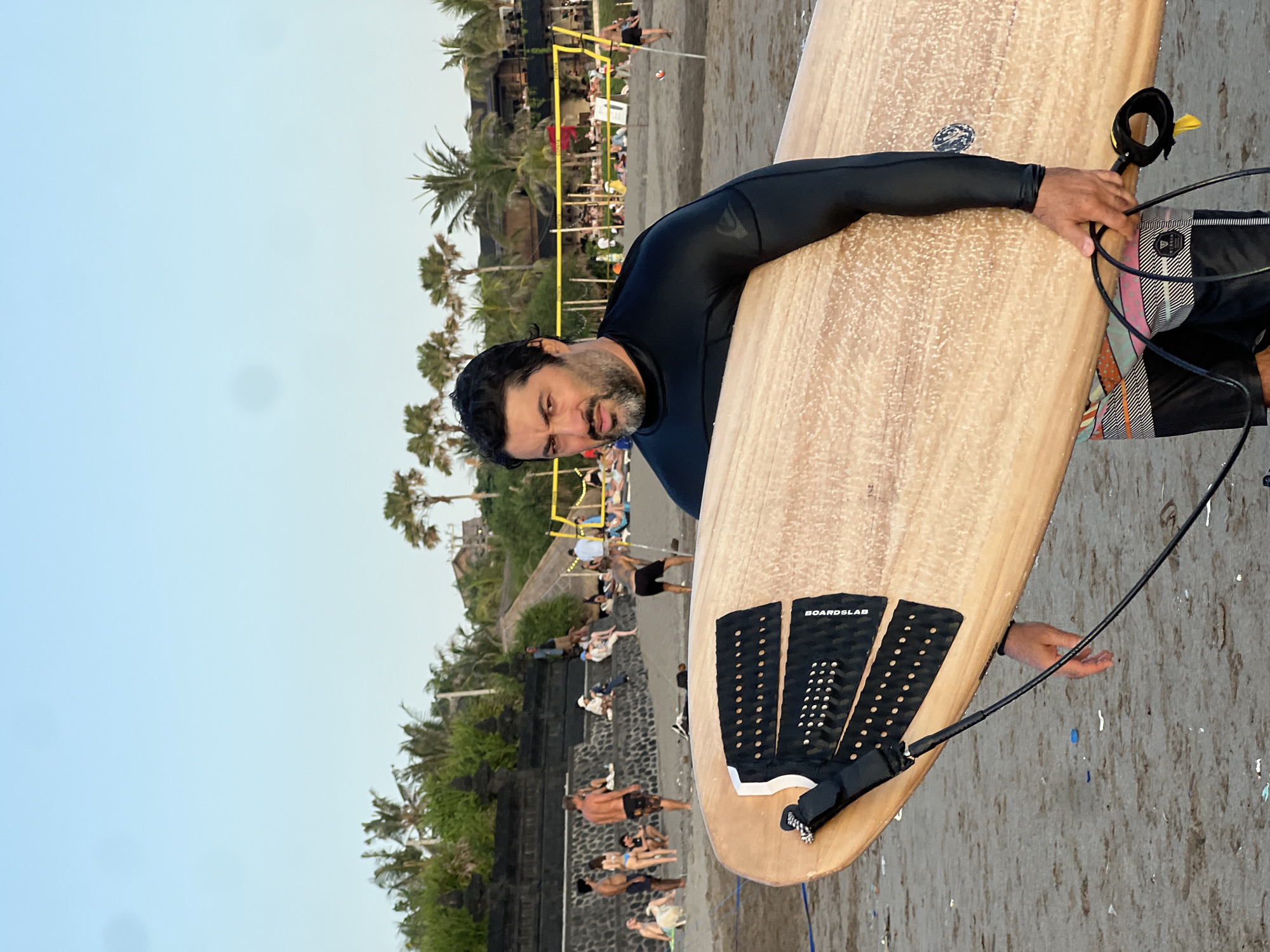 A man in a black wetsuit and patterned shorts carries a light-colored surfboard on a sandy beach in North Kuta, Indonesia. The surfboard is positioned diagonally across his body, held under his right arm and supported by his left hand. His gaze is directed towards the viewer. In the background, palm trees and buildings are visible, with a volleyball net and a scattering of people lounging on the beach. The sky is clear blue.