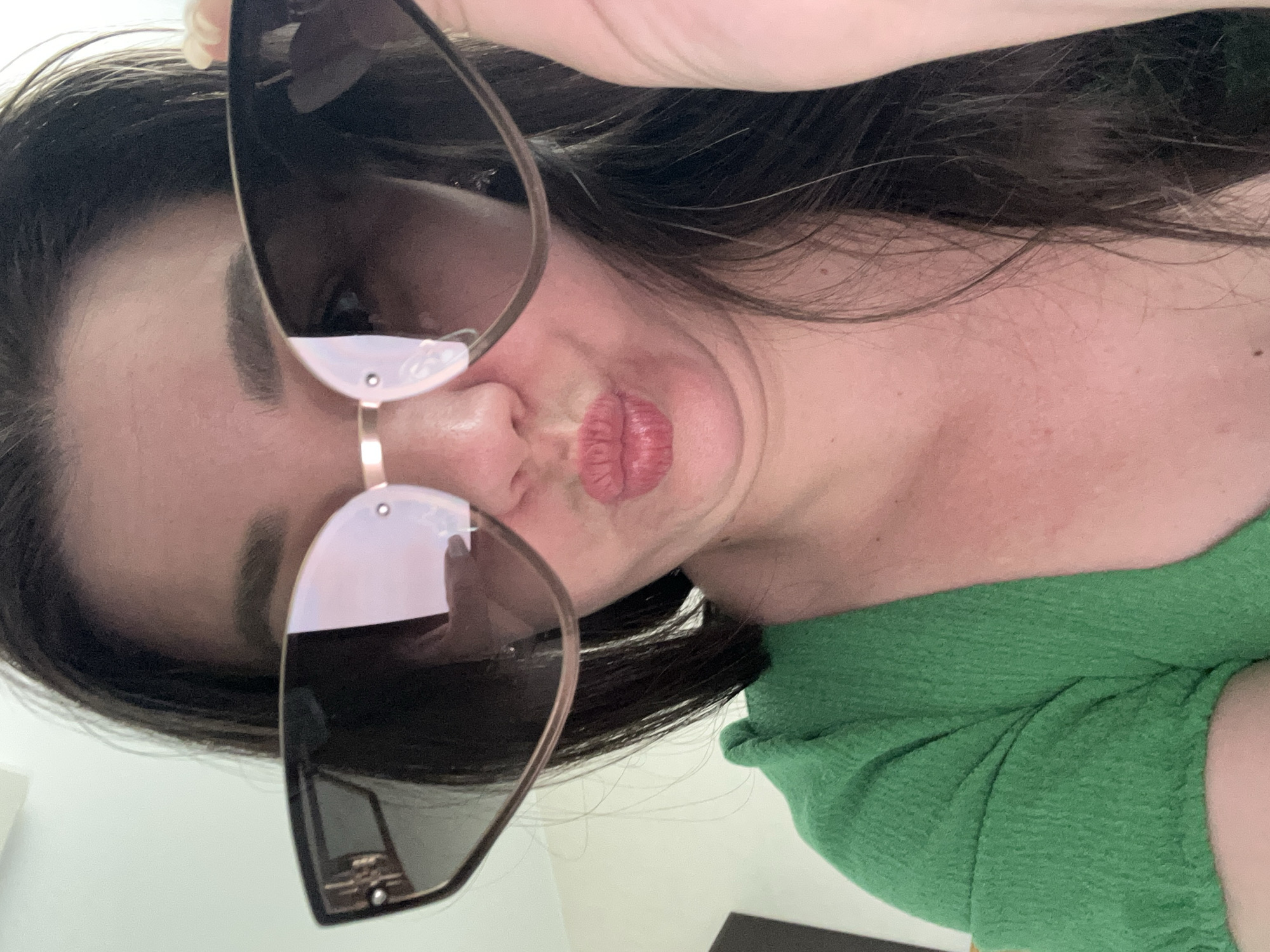 A person with dark hair and fair skin is posing for a selfie. They are wearing sunglasses, which partially obscure their face. They have pursed lips. The person is wearing a green shirt. The background is blurred and indistinct. The location is not discernible from the image, and there is no evidence to suggest the image was taken in Chaikovsky, Russia.