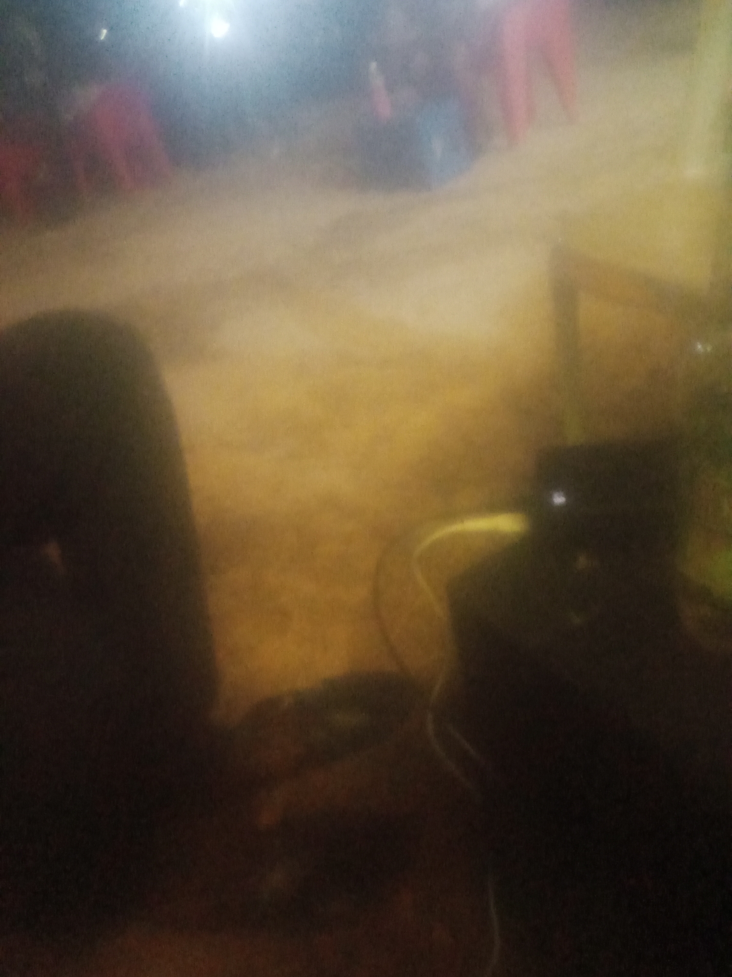 The image is a dark, blurry shot taken at ground level, likely at night. In the foreground on the left, a large, dark, indistinct shape suggests a person or object. To the right, a dark, boxy object with a small white light on its surface is visible, possibly a speaker or electronic device. A light-colored cable snakes across the ground towards it. The background is hazy and illuminated by an orange-yellow light, with faint suggestions of figures and structures, including what appear to be legs in pink and blue. The scene appears to be an outdoor gathering, possibly in a marketplace or street, in Jalingo, Nigeria. The overall impression is one of low light conditions and a lack of clear detail.