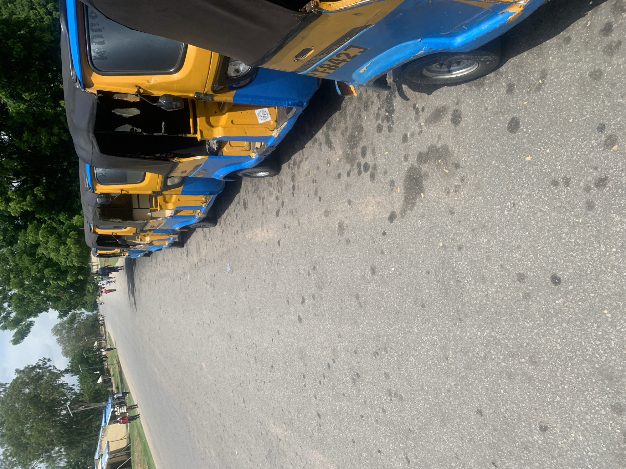 The image shows a street scene in Jalingo, Nigeria, with a row of yellow and blue auto rickshaws parked along the right side of the road. The auto rickshaws are facing away from the camera, towards the distance. Several people are visible on the left side of the road, some walking and others standing near small structures. The road is paved and appears to be a busy thoroughfare. Trees line the left side of the road in the distance, and the sky is overcast. The overall impression is a snapshot of daily life on a street in Jalingo.
