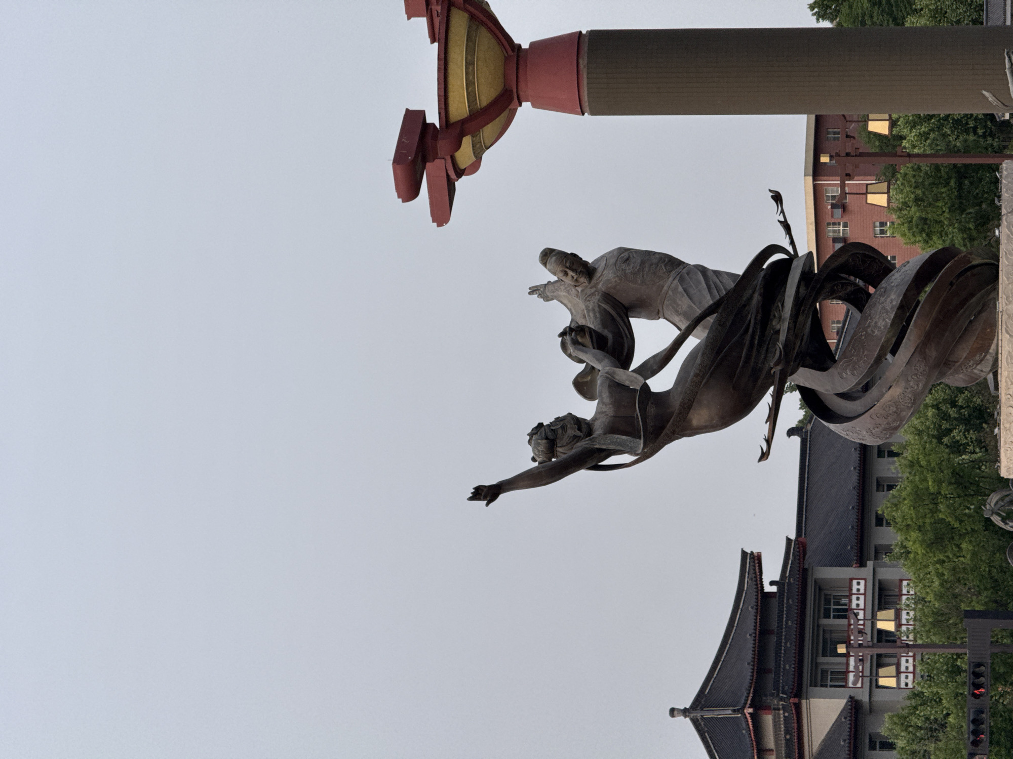 A low-angle shot depicts a bronze statue in Lintong District, China. The statue features two figures enveloped in flowing ribbons, set against a backdrop of a traditional Chinese building with a dark roof and light-colored walls. A red and gold architectural structure stands adjacent to the statue, and the sky is overcast. In the background, glimpses of modern buildings and green foliage are visible. A traffic light pole is present near the base of the building.