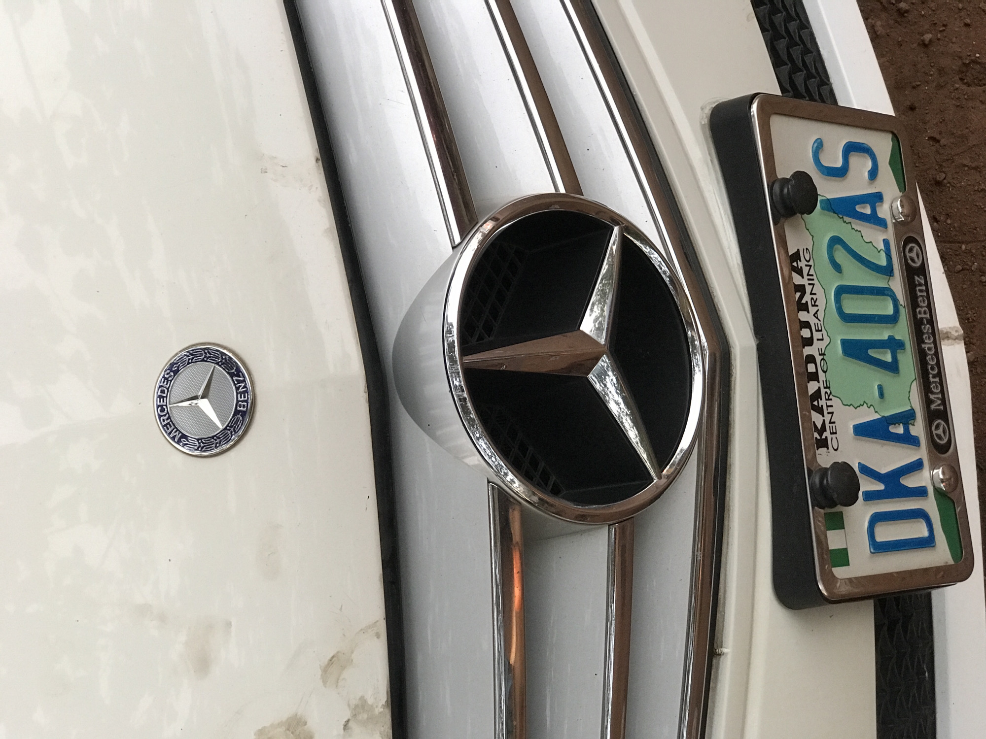 A close-up, high-angle shot captures the front of a white Mercedes-Benz vehicle, focusing on its grille, emblems, and license plate. The car's white paint shows some dirt and smudges, particularly on the left side of the hood.
Dominating the center is a large, chrome Mercedes-Benz star emblem serving as a hood ornament, mounted on the vertical grille bars. To the left, on the flat surface of the hood, is a smaller, circular Mercedes-Benz badge with the brand name "MERCEDES BENZ" and the star logo inscribed within. The grille features several vertical chrome strips set against a lighter, possibly white or silver, background.
On the far right, a license plate is visible, mounted on a black frame. The plate itself has a white background with a green strip running across the bottom and blue characters. The visible text on the license plate includes "KADUNA" (partially visible at the top, likely referring to a Nigerian state), "CENTRE OF LEARNING" (clearly legible), and the registration number "DK-402AS". A small "Mercedes-Benz" logo is also present on the far right of the plate. The immediate surroundings suggest an outdoor setting, with reddish-brown ground visible on the right, consistent with the Galadimawa Village, Nigeria location. The lighting appears soft, indicating it is likely daytime with indirect sunlight.
