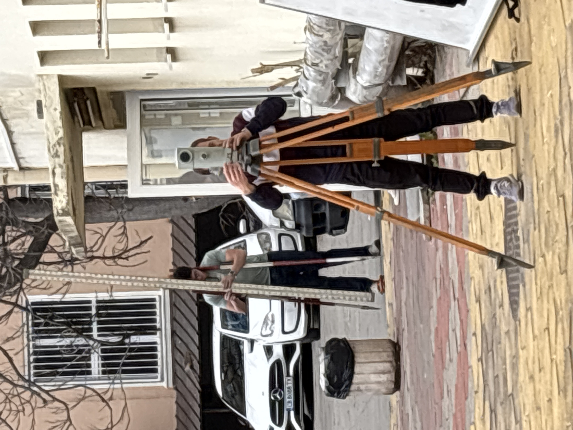 An urban scene depicts two males engaged in land surveying on a paved street in Sofia, Bulgaria.
In the foreground to the right, a male individual with short hair, wearing a dark long-sleeved shirt with white and maroon stripes and black pants, stands operating a grey optical level or theodolite mounted on a wooden tripod. He is looking through the instrument's eyepiece, with both hands positioned on its controls.
In the mid-ground to the left, a second male, wearing a light green short-sleeved shirt, dark jeans, and sandals, holds a tall, telescopic leveling staff vertically with both hands. He is positioned between two parked white vehicles: a Mercedes-Benz with Bulgarian license plate "CB 8088 TX" (indicating Sofia registration) in the foreground, and a smaller Volkswagen Up! behind it.
The immediate ground is paved with multi-colored interlocking pavers, predominantly red and yellow. To the left, a cylindrical waste bin with a black trash bag is present. In the background, a light peach-colored building with a barred window is visible behind bare tree branches, while to the right, a lighter-colored building with recessed vertical features stands, partially obscuring insulated pipes and a white panel. The scene suggests a daytime, outdoor professional surveying operation.