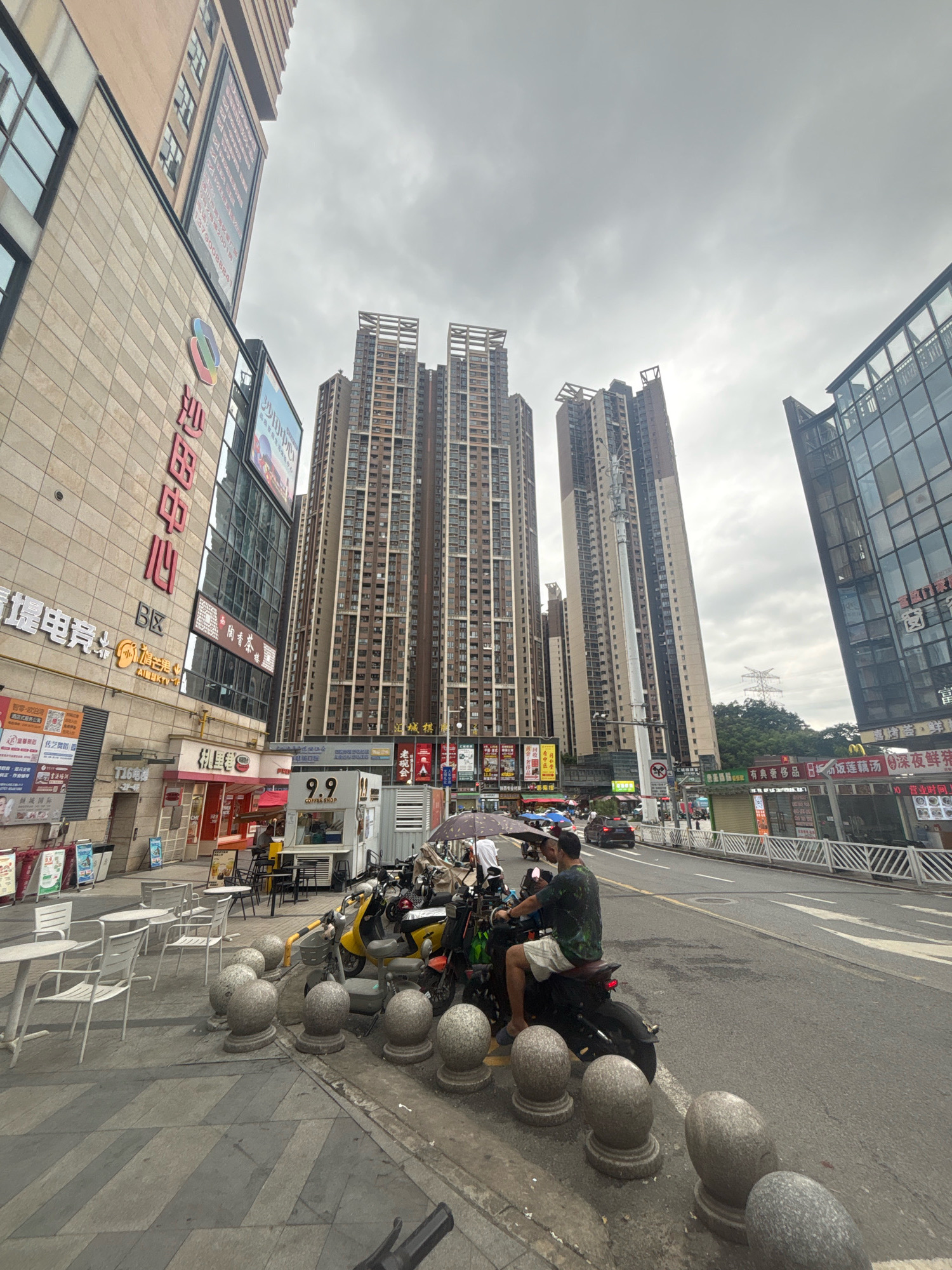 This is a ground-level, wide-angle photograph taken on a cloudy day in Nanhai District, China, likely in a commercial area. The scene is dominated by tall, modern buildings, including a large, light-colored commercial building on the left with red Chinese characters for "Sha Tian Center" visible, and several very tall residential or mixed-use towers in the background.
In the foreground, a paved sidewalk runs along the street. Several white tables and chairs are set up on the sidewalk, suggesting an outdoor dining area. A row of rounded, gray stone bollards separates the sidewalk from the road. A man wearing a green patterned shirt and light shorts is seated on a black electric scooter, facing away from the camera and looking down the street. Other electric scooters and motorcycles are parked nearby. An umbrella is open and positioned over the parked scooters.
The street itself is asphalt with white lane markings and appears to be moderately busy with vehicular traffic, including cars and more scooters. A small kiosk with the number "9.9" and "COFFEE SHOP" is visible on the sidewalk. Advertising banners and signage in Chinese are abundant on the buildings. The sky is overcast, contributing to the diffused lighting. The overall impression is a bustling urban environment with commercial and residential activity.