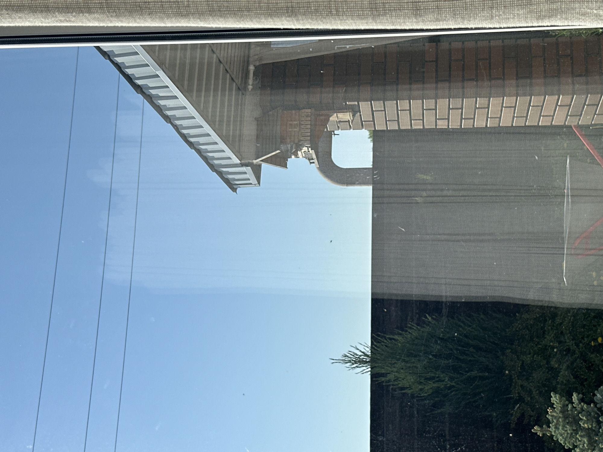 The image depicts a view through a window, split between a clear reflection on the left and a partial view through a screen on the right. The reflection shows a bright blue sky with power lines, and the roofline of a building. The screened area shows the reflection of a brick building with an arched opening and the foliage of green bushes and trees. The window frame and a partially rolled-down shade are visible at the top of the image. Location is unknown.
