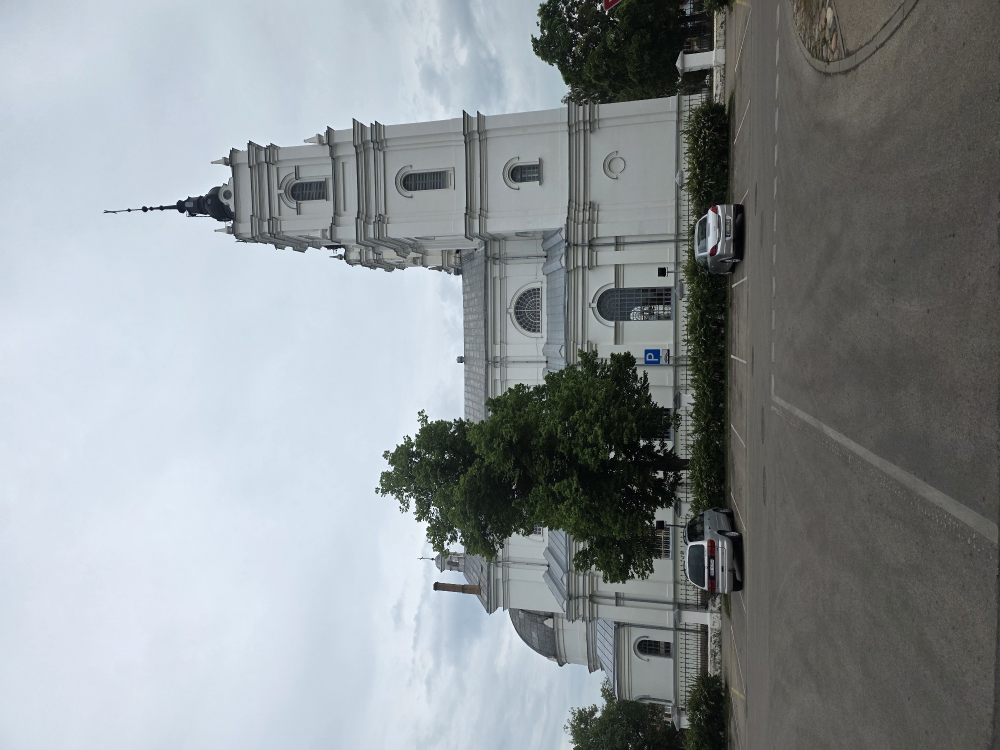 This is a wide outdoor shot of a large, imposing white church or cathedral under an overcast sky. The building features a prominent, multi-tiered bell tower on its right side, capped with a black dome, a slender spire, and a cross. The church facade is uniformly white, adorned with various arched, rectangular, and semi-circular windows, and grey sloped roofs are visible above its multiple sections. A smaller, grey dome is present on the left side of the structure, partially obscured by a large, leafy green tree.
In the foreground, a grey asphalt parking lot occupies the lower half of the image. White lines delineate parking spaces. Two silver cars are parked in the lot: a minivan (license plate GQL 934) on the left and a sedan (license plate VDE 731) on the right. A low black metal fence, interspersed with white stone pillars, separates the parking area from the church grounds, which are lined with green bushes. A blue parking sign with a white 'P' is visible near the fence on the left. The sky is a uniform, light grey, indicative of heavy cloud cover, and no individuals are present in the scene.