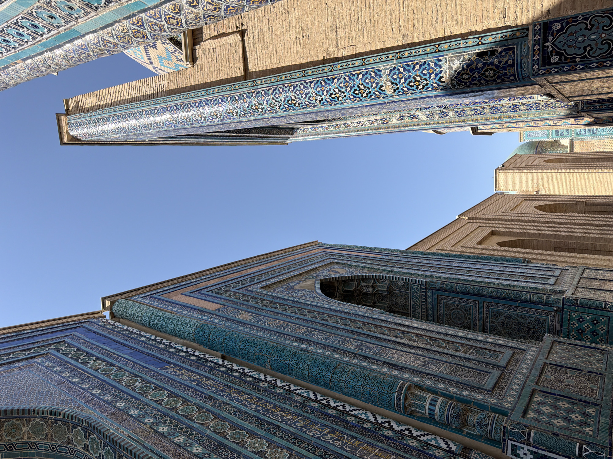 This is a low-angle, full-shot, eye-level, exterior, daytime photograph of the Shah-i-Zinda necropolis in Samarkand, Uzbekistan. The buildings are constructed of brick and adorned with intricate blue and turquoise tilework. Two towering structures with a variety of geometric and floral tile patterns dominate the foreground, leaning inwards towards the center of the frame. The building on the left features a large, recessed archway. The building on the right is a tall, slender pillar also covered in detailed tiling. Behind these structures, more brick buildings with arched openings are visible, along with a distant turquoise dome. The sky is clear and blue. There are no people visible in the image.