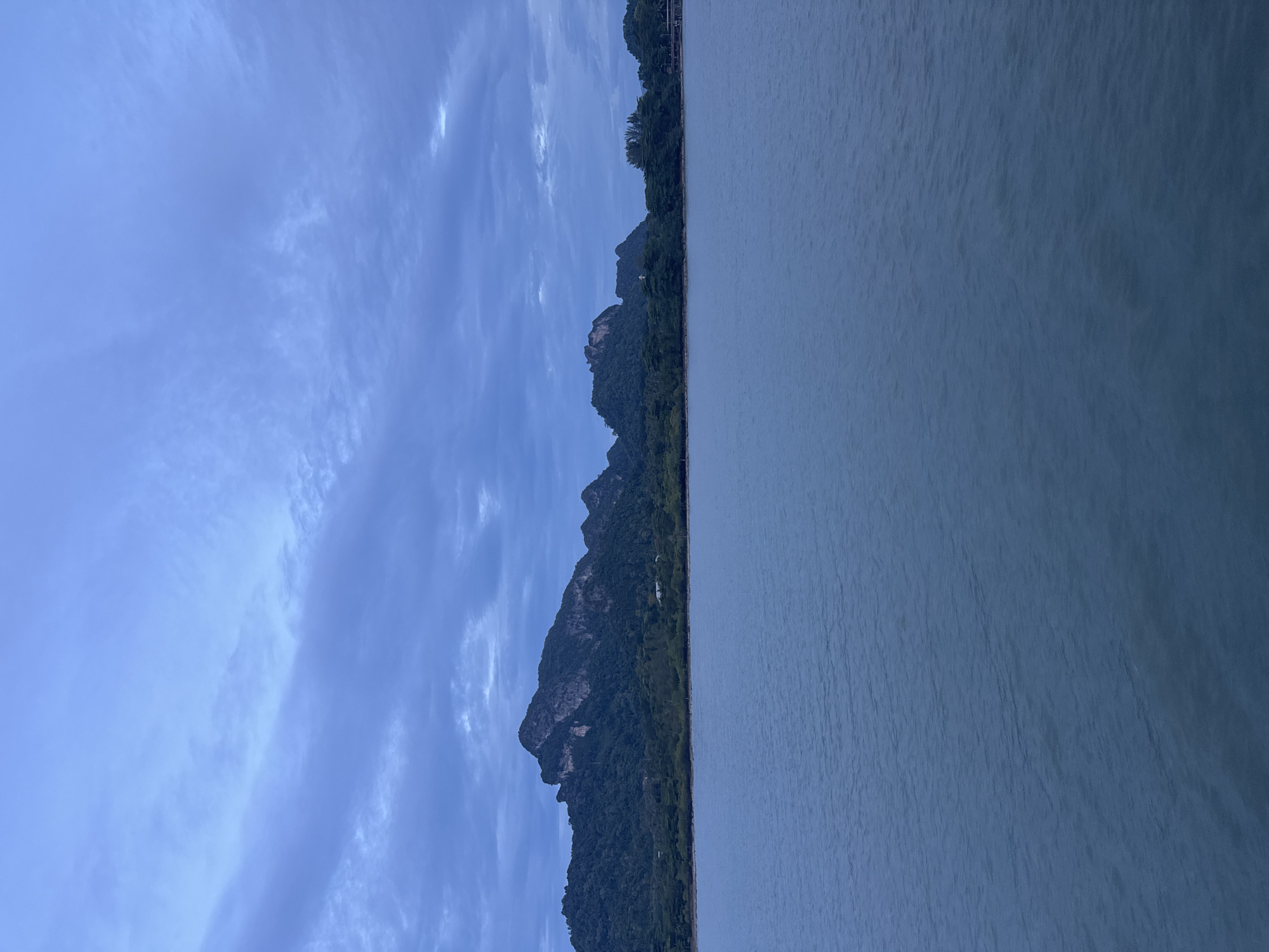 This image depicts a serene natural landscape scene. The foreground is dominated by a calm body of water, likely a sea or large lake, its surface reflecting the muted tones of the sky. The water appears to be a deep, tranquil blue, with subtle ripples suggesting a gentle breeze.
In the midground, a lush, green coastline rises dramatically, characterized by steep, forested mountains. The dense foliage covers the slopes, with the rugged rock formations of the peaks visible through the trees. A thin strip of land runs along the base of the mountains, where a few scattered structures or buildings can be glimpsed amidst the greenery. The overall impression of the land is one of wild, untamed nature.
The sky above is a canvas of soft blues and grays, with delicate, wispy clouds stretching across the expanse. The lighting suggests it is either early morning or late evening, as the sun is not directly visible and the light is diffused. The atmosphere is calm and peaceful, evoking a sense of quietude and natural beauty.
The composition is split horizontally, with the water occupying the right half of the frame and the land and sky the left. This division emphasizes the vastness of both the water and the sky in contrast to the solid mass of the mountains. There is no visible text in the image. The scene appears to be a natural and undeveloped location, possibly a coastal or lakeside area.