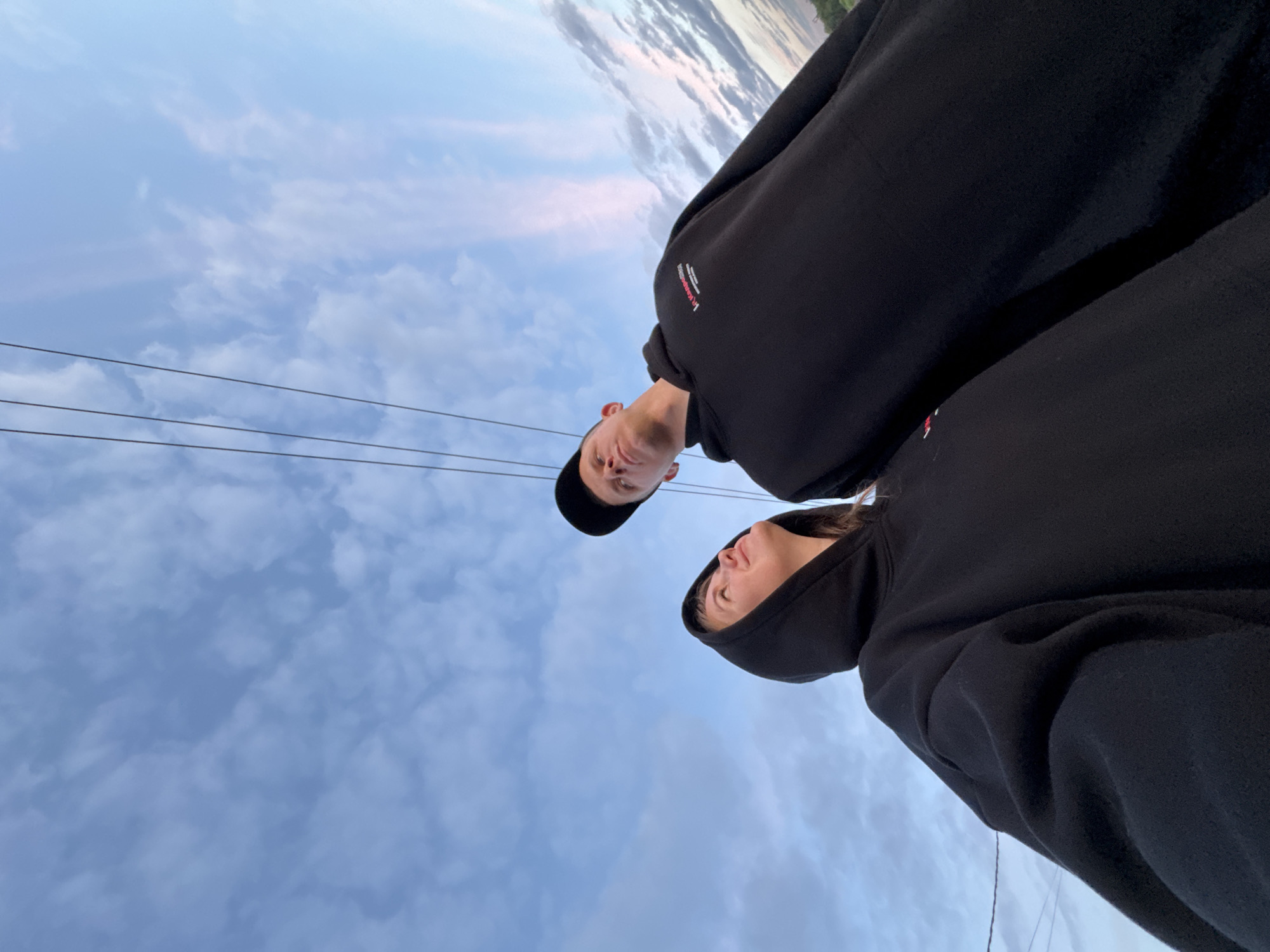 Two individuals, a man and a woman, stand close together, looking upwards towards the sky. Both are wearing black hoodies. The man is also wearing a black cap. The sky is visible in the background and consists of blue and white clouds. Overhead power lines are visible crossing the sky. The location appears to be in Горный, Russia.