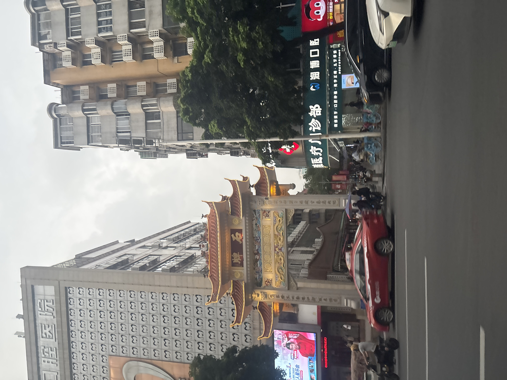 The image captures a street scene in Tianhe District, China. Dominating the left side is a multi-story building with a sign at the top displaying Chinese characters that appear to be "口腔医院" (Dental Hospital). The facade of this building is decorated with a repeating pattern of what looks like stylized teeth or mouths. To the right of the "口腔医院" building, a traditional Chinese archway, likely a paifang, stands prominently. This archway is elaborately decorated with golden embellishments and features Chinese characters on its central beam, reading "牌石" (Stone Tablet). Above the archway are layered, curved roofs with ornate detailing.
In front of the archway, a large digital billboard displays an image of a man in a red traditional outfit, possibly promoting something. Further to the right, another building facade features a bright teal sign with white Chinese characters, including "医疗门诊部" (Medical Outpatient Department) and "口腔" (Dental). This building also has a red logo.
On the street, a red sedan car with white racing stripes is a central focus, positioned in the foreground. A person on a small, black motorcycle is seen to the left of the red car, moving in the same direction. In the background, several other vehicles, including a black car and a white car, are visible, as are bicycles parked on the side of the street. People are also seen walking and standing on the sidewalk. The overall environment suggests a busy urban area with a mix of modern and traditional architecture. The sky is overcast, indicating a cloudy day.