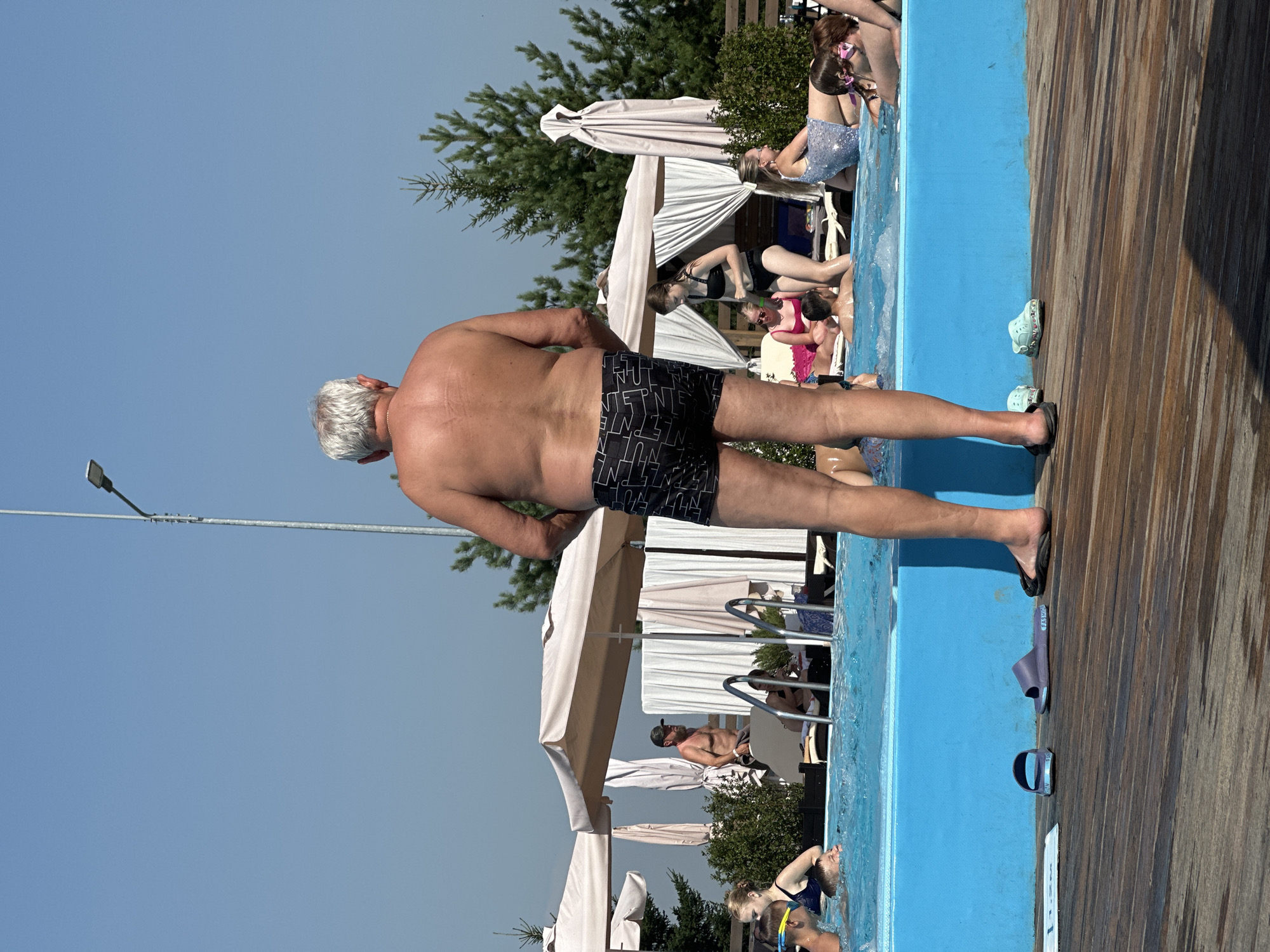 The image depicts a poolside scene on a sunny day. A shirtless, elderly man with grey hair stands facing away from the camera, wearing patterned swim trunks. In the background, a pool with bright blue water is populated by several people. Some are wading, while others are relaxing on lounge chairs under canopies. A lamppost is visible against a clear blue sky, along with evergreen trees. The foreground shows a wooden deck with several pairs of sandals and a depth marker indicating "1.2M". The overall atmosphere suggests a public recreational area, likely an outdoor swimming pool.