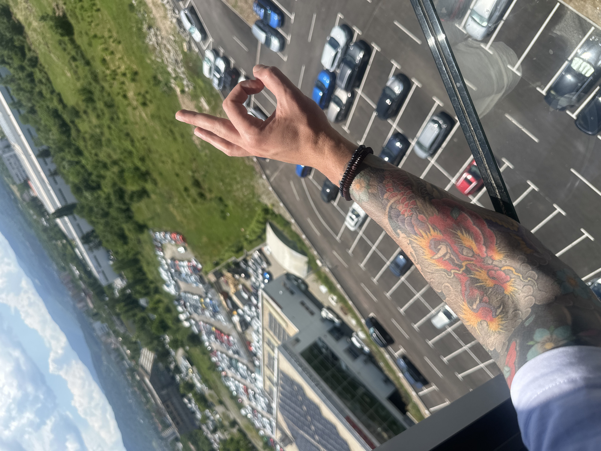 The image features a person's arm with a prominent tattoo of a dragon and floral motifs, positioned in the foreground. The arm is extended, with the hand making a gesture where the thumb and index finger form a circle. The background depicts a wide view from a high vantage point, overlooking a parking lot filled with numerous cars. Beyond the parking lot, there is a green, grassy area and buildings. The scene suggests a bright, possibly sunny day, given the lighting. The perspective is from above, looking down at the cars and the surrounding landscape, which includes hints of a city with distant mountains or hills visible under a partly cloudy sky. The overall impression is one of a casual, possibly reflective moment captured from a significant height, with the detailed tattoo adding a personal and artistic element to the scene. There is no visible text in the image.
