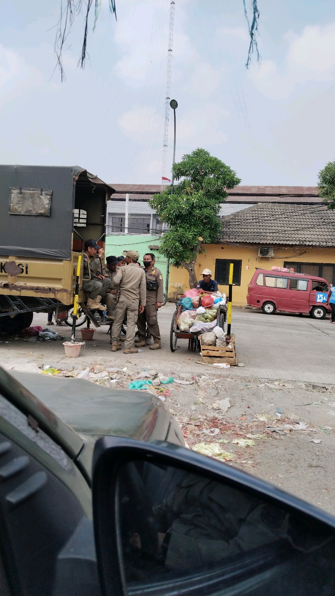 In the city of Bekasi, Indonesia, a group of individuals, appearing to be law enforcement or security personnel in uniform, are gathered near a parked military-style truck. One individual is seated on the truck bed, while others stand and converse. A vendor's cart laden with produce, including watermelons and various bagged items, is positioned nearby. A red minivan is also visible in the background, along with a tall communication tower. The ground is littered with debris and trash, indicating an urban environment that may be experiencing sanitation challenges. The sky is overcast with some clouds. The scene suggests a potentially routine interaction or a pause in an operation.