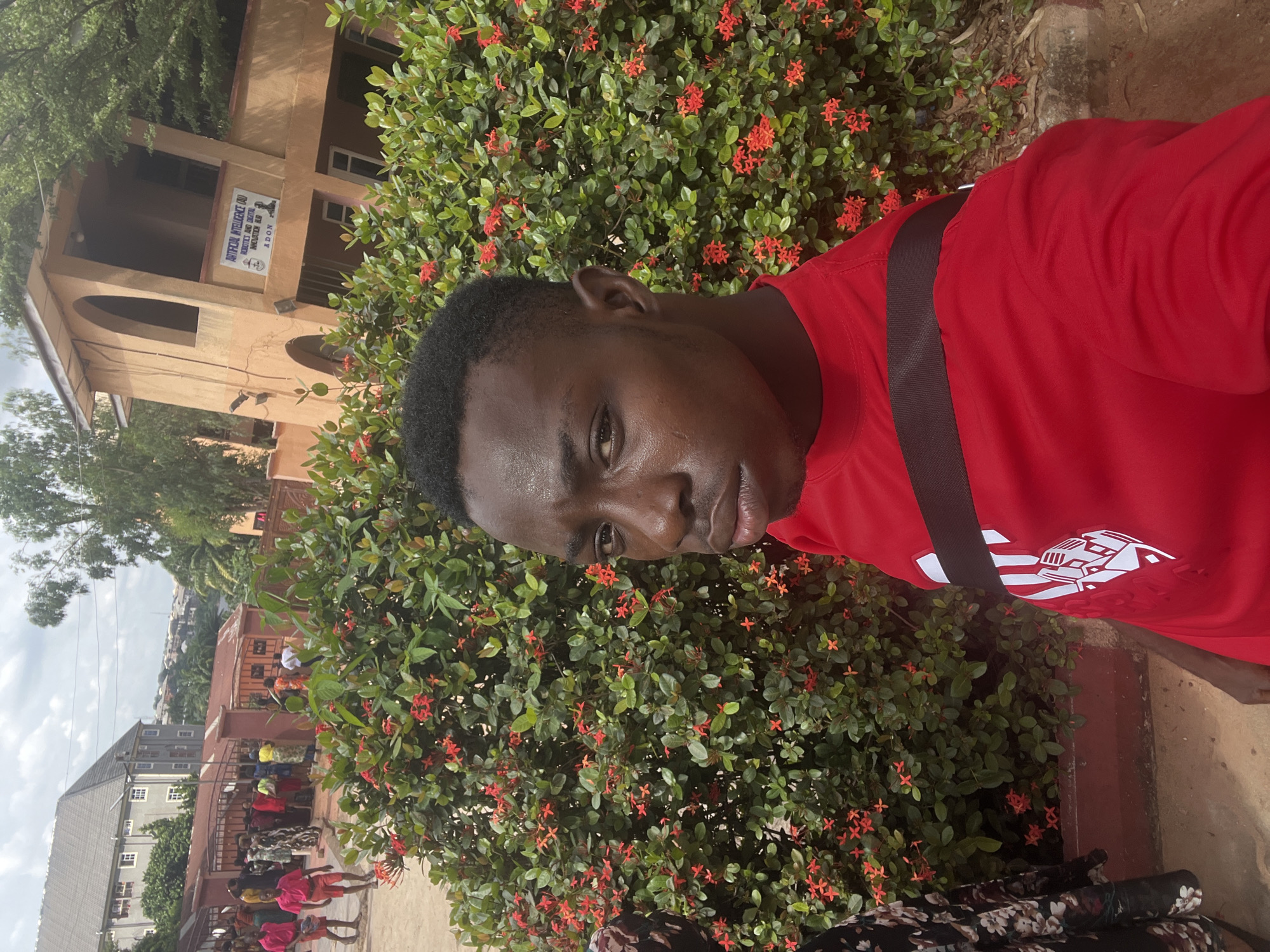 A young Black male in a red t-shirt with a black strap across it poses for a selfie in front of a large green bush with small red flowers. He is in the foreground, and the background shows a street with other people and buildings. One building in the background has a sign that reads "ARTIFICIAL INTELLIGENCE (AI) ROBOTICS AND DIGITAL INNOVATION HUB ADON." The scene appears to be located in Nnewi, Nigeria. The sky is overcast.