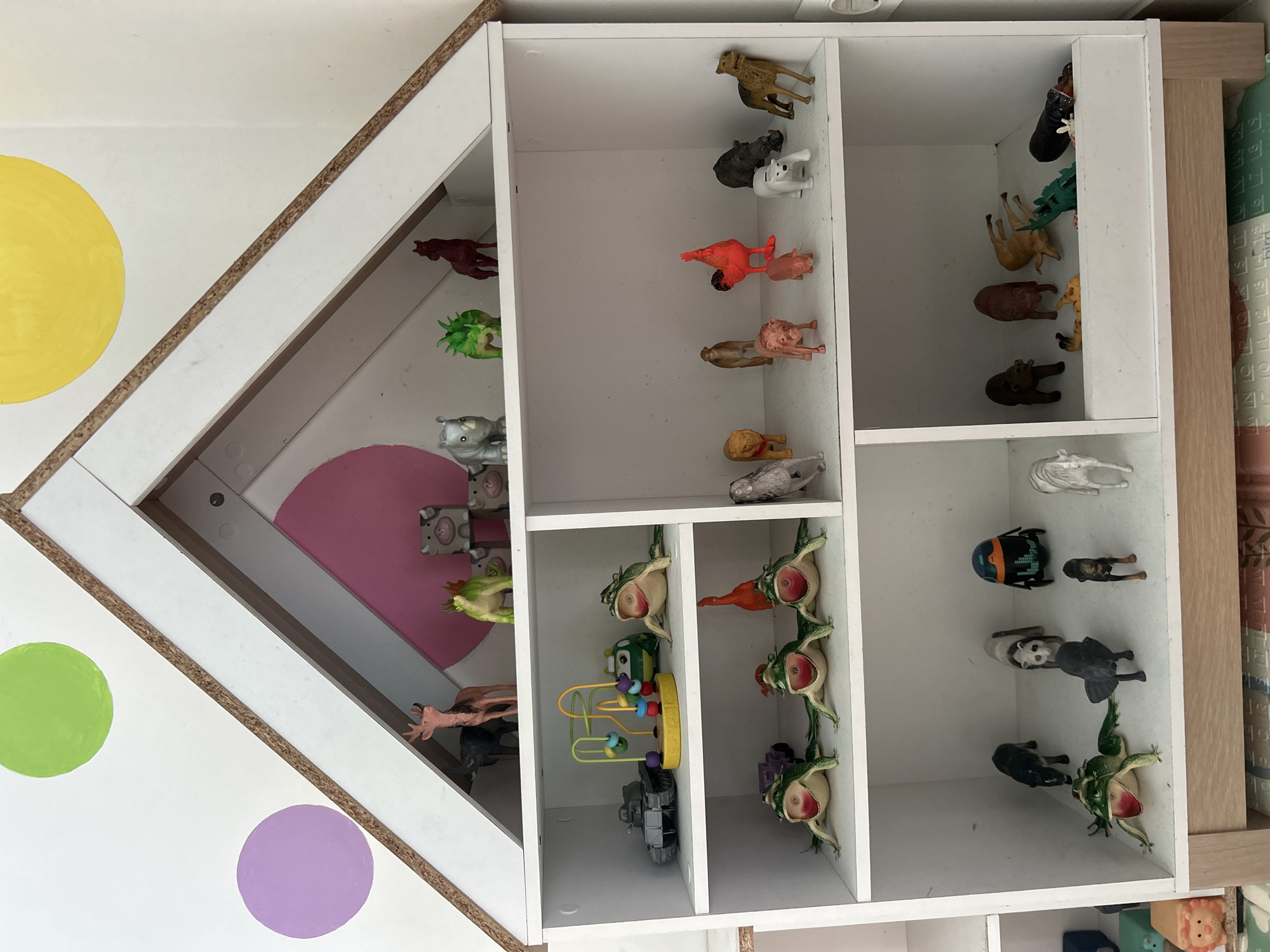 A white, two-story toy shelf in the shape of a house is filled with various toy animals and a few vehicles. The background is a white wall with large, colorful circles: a green one on the upper left, a purple one below it, and a yellow one on the upper right. The shelf is positioned indoors.
On the top shelf of the house-shaped part of the shelf, there is a brown deer with antlers, a black elephant, a green dinosaur-like creature, and a dark brown horse. To the right, on the main shelf structure, there are three grey cow heads stacked, and to their left, a grey rhinoceros.
The second shelf from the top has a grey toy tank on the left, followed by a colorful bead maze toy. On the right side of this shelf, there are three green frogs with their mouths open and red insides, and to the far right, a grey animal that appears to be a seal or a whale.
The third shelf has three more green frogs lined up horizontally across the left side, with a purple toy car behind the first frog. On the right side of this shelf, there is a small orange rooster, a pink pig, a light brown camel, a tan lion, a grey animal that looks like a wolf or fox, and a white polar bear. Further to the right, there is a dark grey rhinoceros and a tan dog with its head turned to the right.
The bottom shelf is split into two sections. The left section contains a black horse, a green frog with its mouth open, a black dog, and a white dog that appears to be a poodle. On the right side of the bottom shelf, there are more toy animals including a brown bear, a brown animal that looks like a deer or cow, a tan animal with its legs in the air, a green dinosaur, and a brown animal lying down.
The toy shelf is situated in a room in Kuta, Indonesia.
