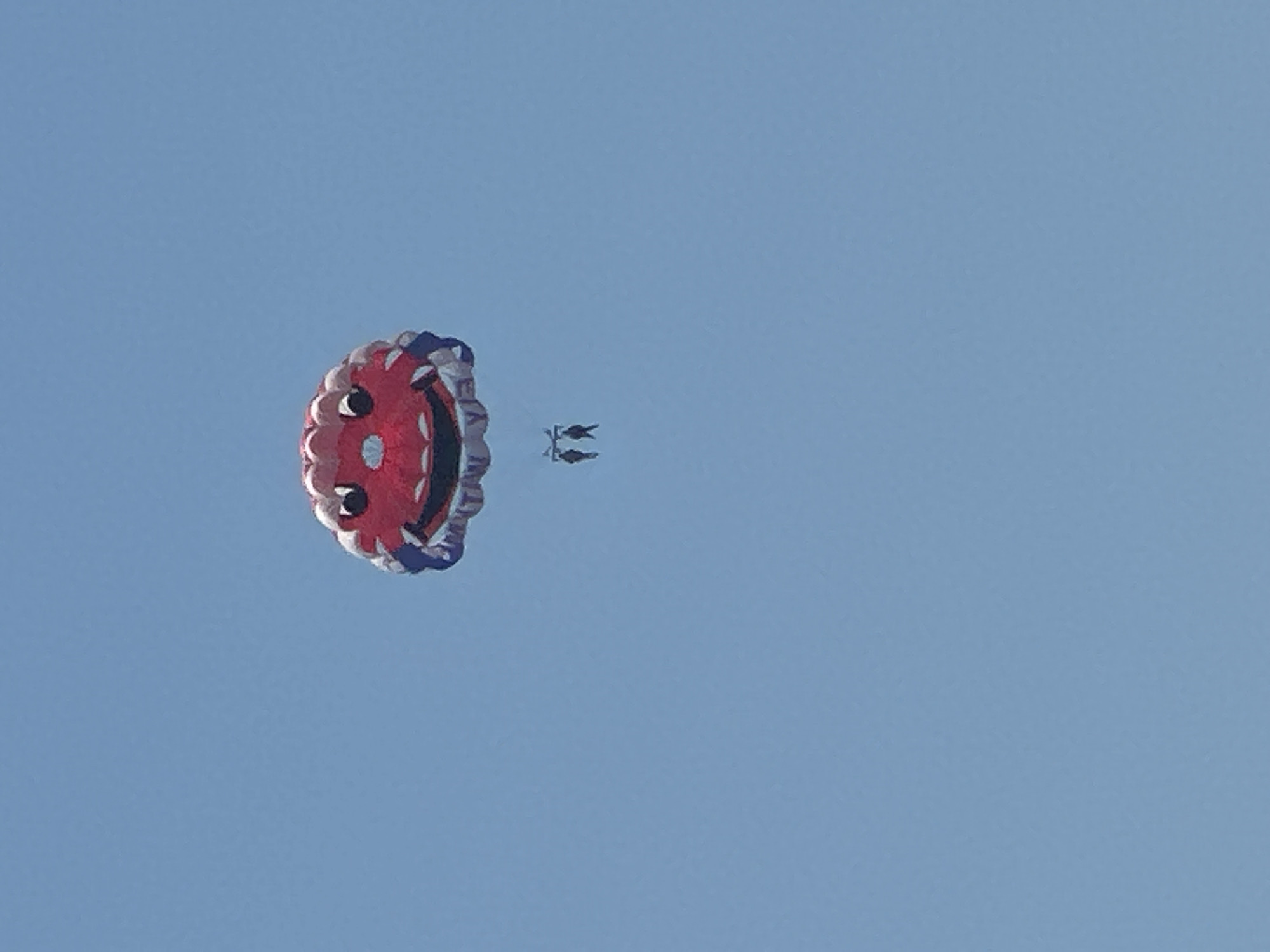 An overhead view shows a parachute, shaped and colored like a smiling red face with blue and white trim, against a clear, light blue sky. Two small figures, appearing to be people, are suspended beneath the parachute by ropes. The scene appears to be captured from below, looking upwards. The image is taken in Budva, Montenegro, as indicated by the presence of this paragliding activity over the city.