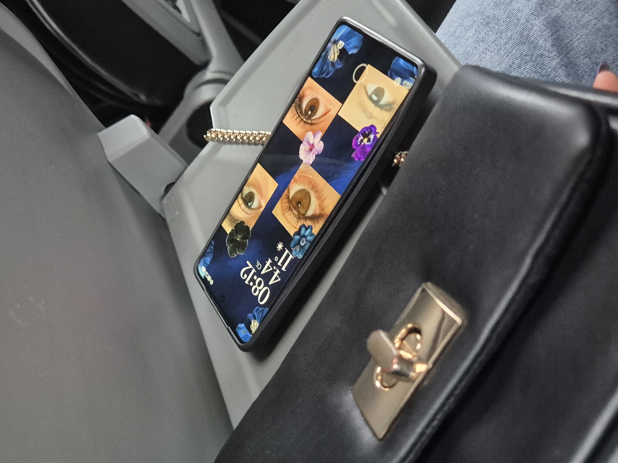 A phone displays a background of eyes and flowers, showing a time of 08:12 and 11.4 degrees, situated near a black purse with a gold clasp. The location appears to be indoors within a vehicle. The specific city of Peichinovo, Bulgaria is not discernible from the image.