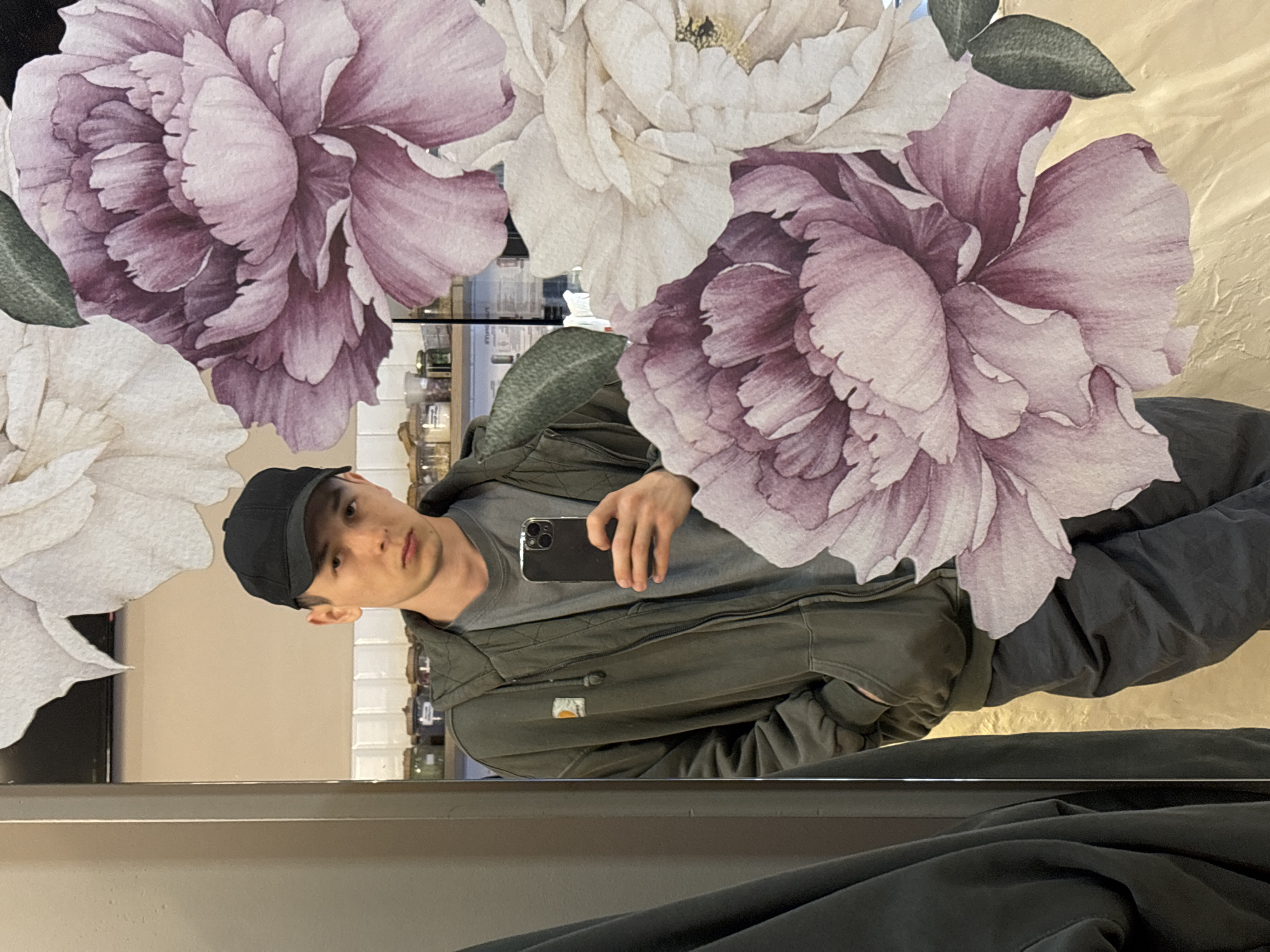 A young man, wearing a black baseball cap, a grey t-shirt, and a dark green zip-up hoodie, stands in front of a mirror in what appears to be a retail or home environment. The mirror reflects the man and the background behind him. The background shows shelves stocked with various jars and containers. A large floral decal, featuring pink and white peonies, is prominently displayed on the mirror, partially obscuring the reflection. The man is holding a smartphone in his right hand, as if taking a selfie. He is wearing dark grey pants. The overall scene suggests a casual moment captured by the individual. The location of Misaylovo, Russia is not evident from the visual information provided in the image.