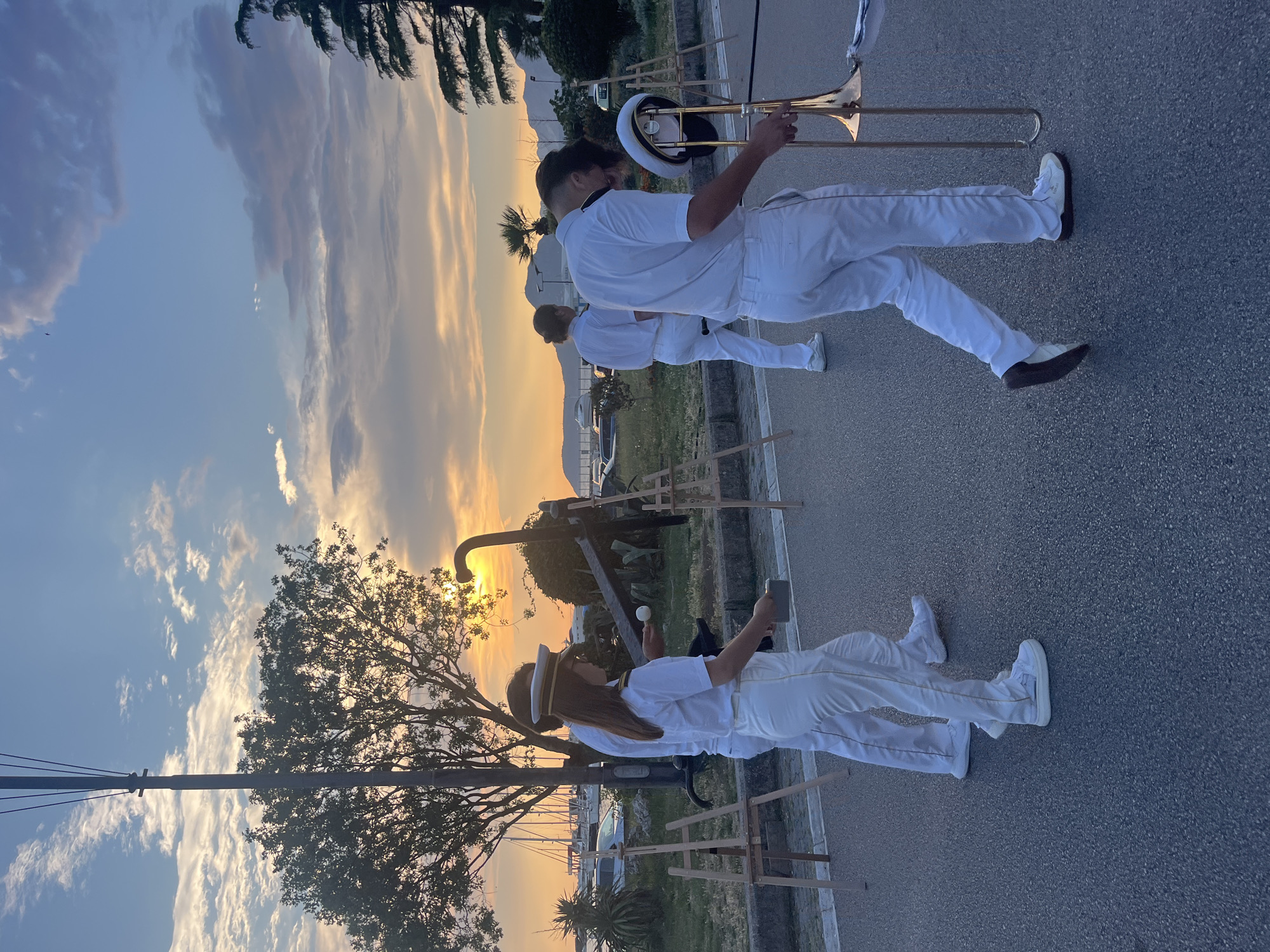 In Šušanj, Montenegro, at sunset, a group of individuals in white uniforms, possibly a marching band, are walking on a paved road. One person in the foreground is holding and walking with a trombone. Another individual in a white uniform and hat is walking with a small drumstick and what appears to be a small drum or percussion instrument. In the background, there are trees, yachts in a harbor, and mountains. The sky is filled with dramatic clouds tinged with the warm colors of the setting sun. Several easels are set up along the side of the road.