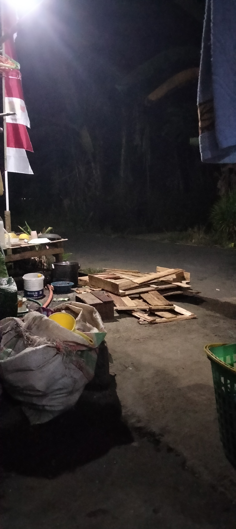 The image depicts an outdoor scene at night in Serut, Indonesia. In the foreground, a large, loosely woven sack is partially visible, containing a yellow funnel and other indistinct items. To the right of the sack, a green plastic basket is partly visible. Behind the sack, a pile of wooden planks and other lumber is stacked on the ground. Further back, a wooden table is set up with various items including a white bucket, a blue bowl, and a hose. A flag with red and white stripes is attached to a pole on the left side of the scene, illuminated by a bright light source from above. The background consists of a dark, indistinct area with trees and vegetation, suggesting a rural or semi-rural setting. The ground appears to be asphalt or packed dirt. The overall impression is one of an outdoor workspace or storage area at night.