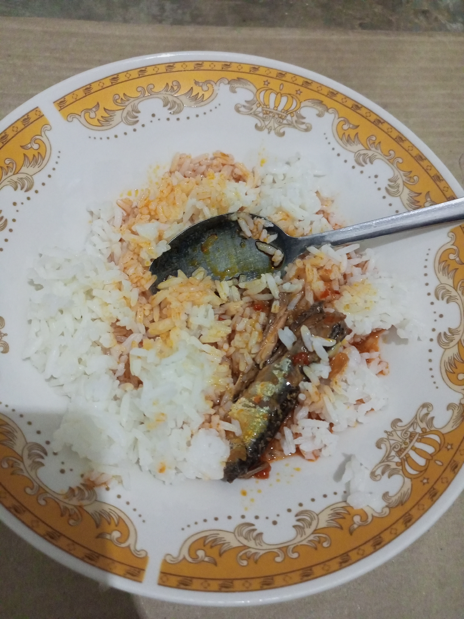 A close-up, eye-level shot shows a plate of rice with sardines in sauce. The plate has a decorative gold rim with a crown motif. A silver spoon is resting on the rice, partially submerged. The background is a blurry, beige surface that appears to be a table. There is no visible indication of the specific location in Batam, Indonesia.