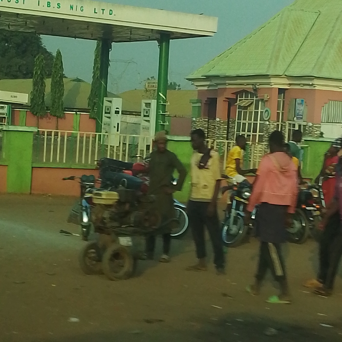 A group of people congregates near a gas station with the sign "JOST I.B.S NIG LTD." above. Several motorcycles are visible, along with a small, wheeled contraption near the foreground. The building beside the gas station is a peach color with a green roof. A man in an olive green outfit stands with his hands on his hips. Other people in the group are wearing yellow and pink jackets. The setting seems to be a street or open area near the gas station.