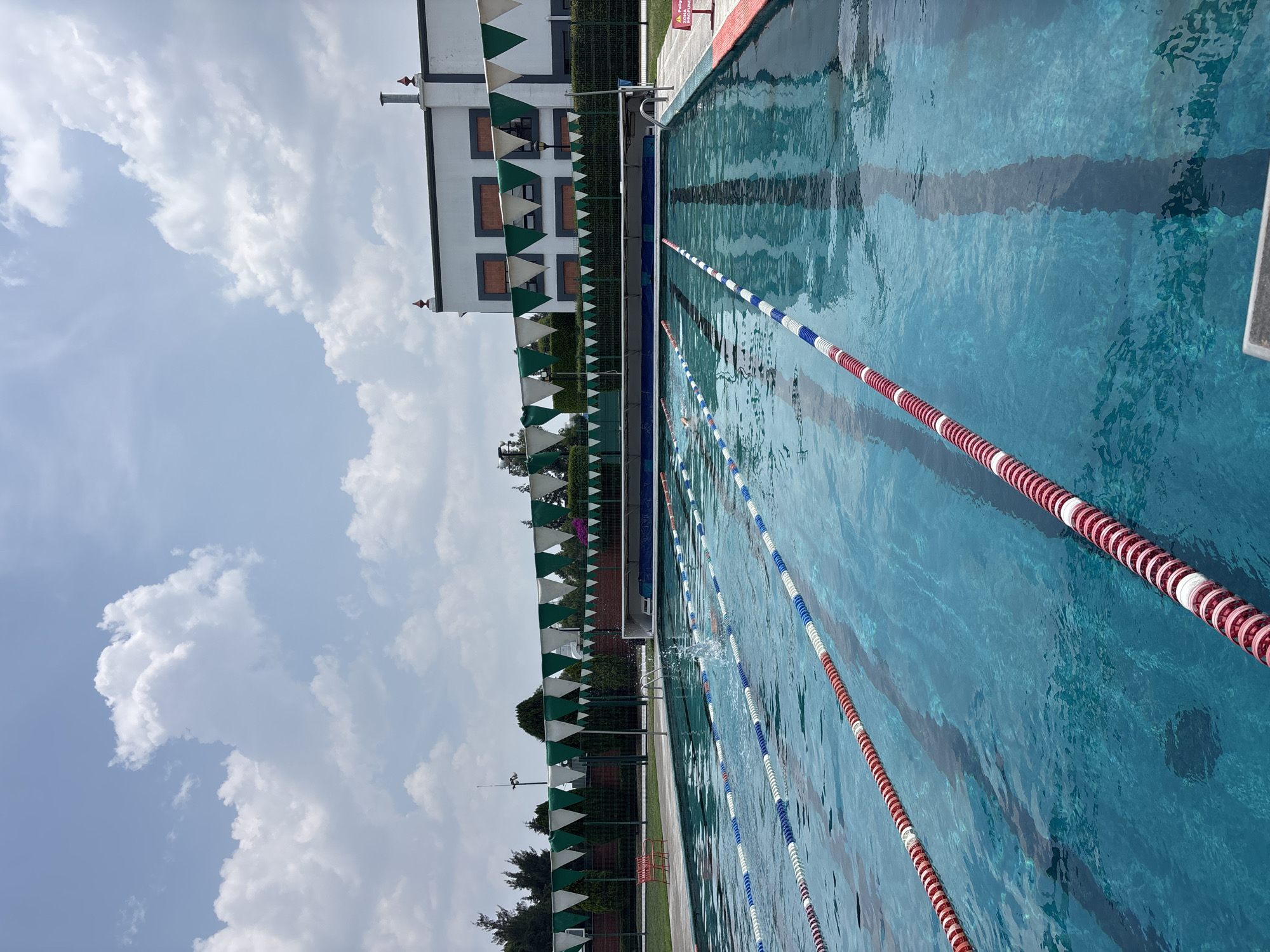 The image shows an outdoor swimming pool located in San Andrés Cholula, Mexico. The pool is divided into lanes by floating lane markers, alternating between red/white and blue/white color schemes. The water is clear and blue, with reflections visible on the surface.
Along the poolside, there's a fence adorned with triangular pennants in green and white. Beyond the fence is a building with a white facade and dark grey accents, punctuated by windows. The sky is partly cloudy.