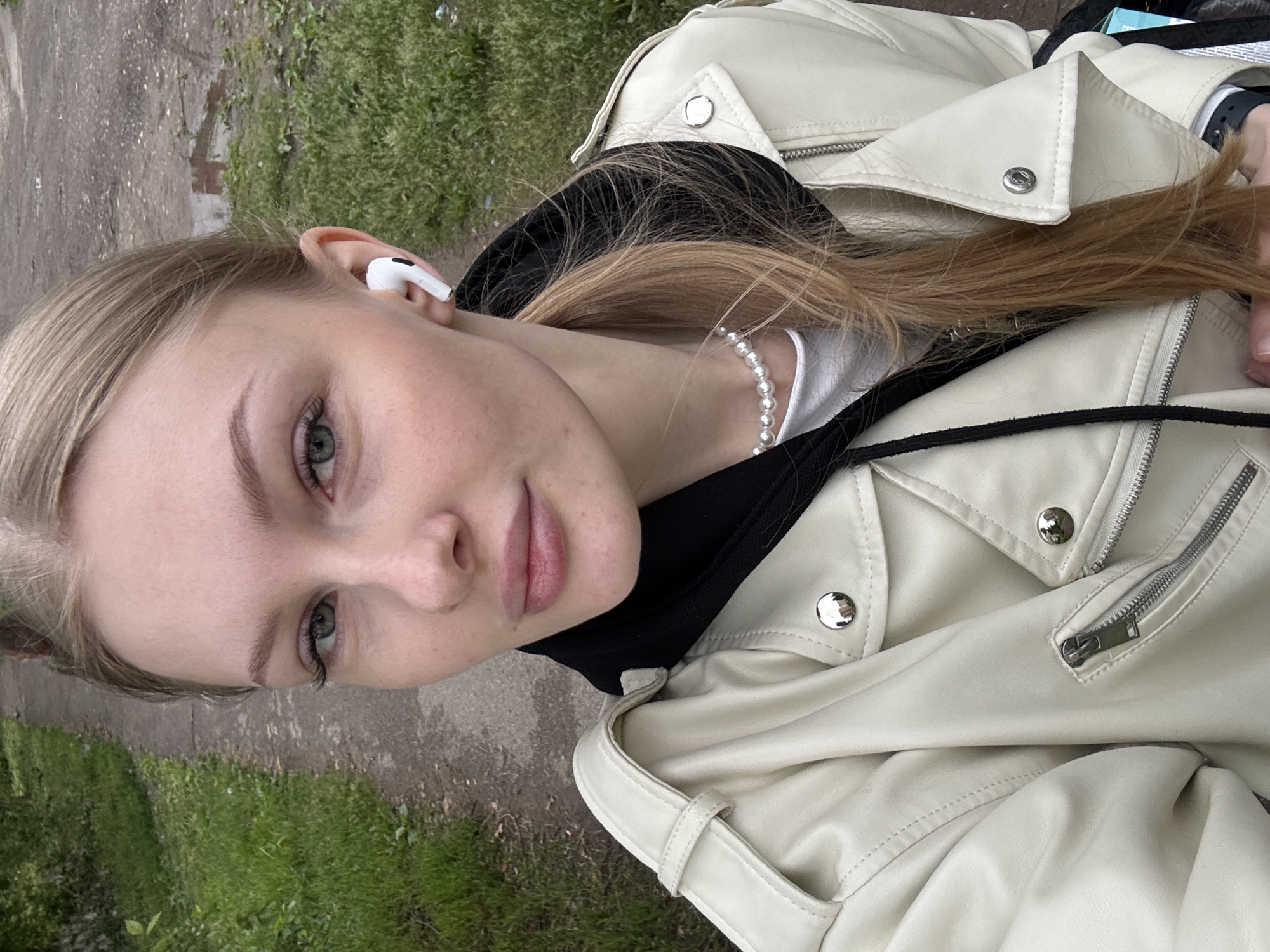 A young adult female is depicted in a close-up, chest-up self-portrait taken outdoors in Perm, Russia. She has a fair complexion, light-colored eyes (appearing greenish-blue), and light blonde/light brown hair pulled back into a ponytail, with some strands loose over her right shoulder and around her face. Her eyebrows are well-defined and light in color. She has a subtle, closed-mouth smile.
She is wearing an off-white or cream-colored faux leather moto-style jacket with silver zippers and snap buttons, including visible lapels and a zippered chest pocket on the wearer's left side. Beneath the jacket, a black hooded sweatshirt is visible, with its hood forming a collar around her neck and black drawstrings visible. A white crew-neck top is worn underneath the hoodie. A white pearl choker necklace is visible around her neck, above the white top. A white Apple AirPods Pro earbud is securely placed in her right ear. Part of a dark-colored smartwatch or fitness tracker is visible on her left wrist.
The background features an outdoor path, appearing to be concrete or asphalt, which is somewhat uneven and worn, extending into the distance. Green grass and various foliage line both sides of the path, with denser vegetation on the left side suggesting a treeline or shrubbery. On the right side of the path, patches of dirt and small puddles of water are discernible, indicating either recent precipitation or generally unpaved sections. The lighting is overcast and diffused, suggesting a cloudy day.