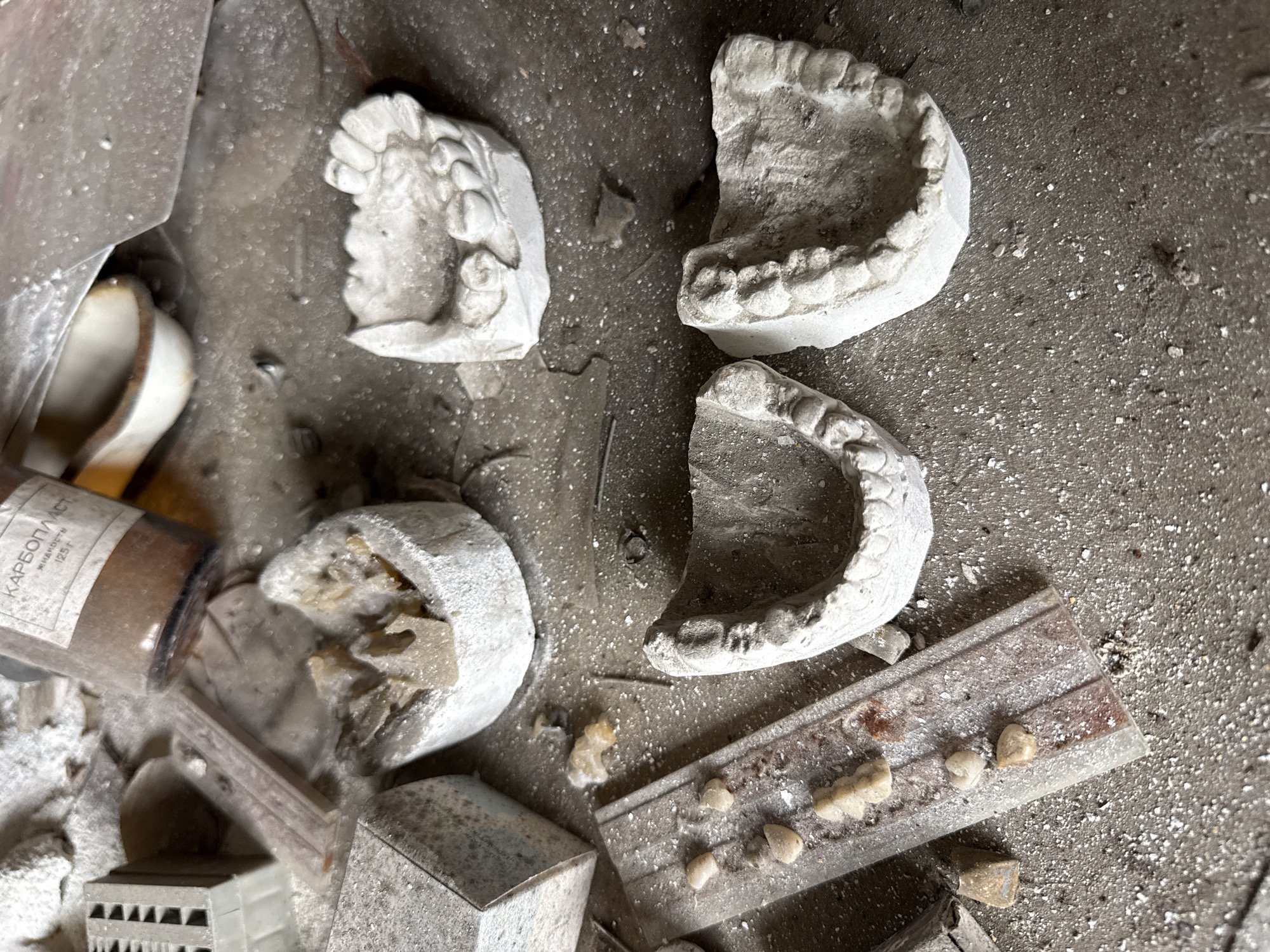 The image depicts a scene in what appears to be an abandoned dental office or laboratory in Prypiat, Ukraine. Four plaster dental molds lie broken on a dusty, debris-strewn floor. A tipped-over bottle labeled "Карболист" (Carbolist) is partially visible, suggesting a chemical solution formerly used in the space. A segmented ruler rests among fragments of other objects, possibly architectural models or building components. The environment is characterized by decay and neglect, consistent with the abandonment of the city after the Chernobyl disaster. There are no people visible in the scene. The overall impression is of a space abruptly left behind, with the tools and artifacts of a profession abandoned mid-use.