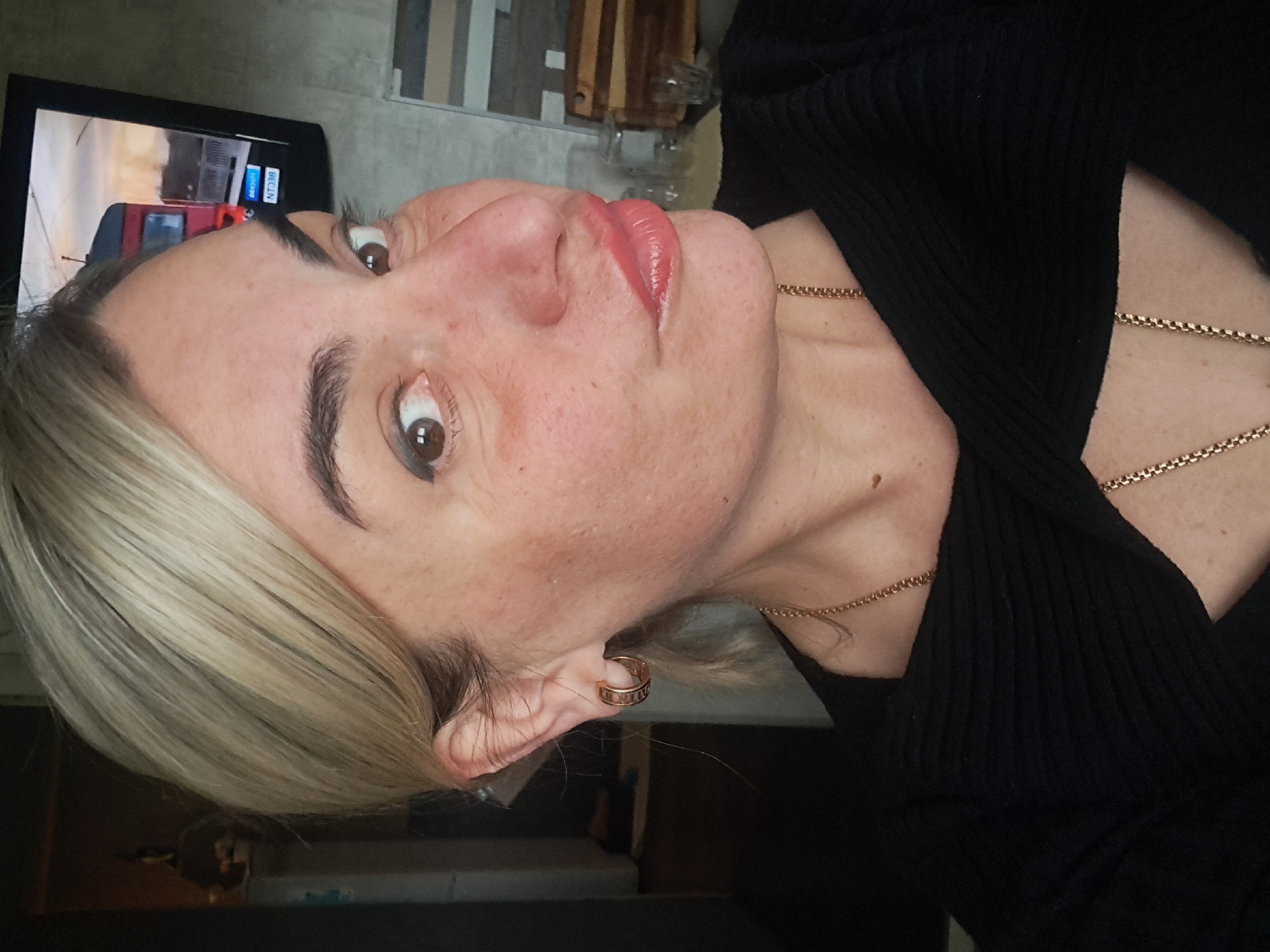 A woman with blonde hair is facing the camera, wearing a black top and two gold necklaces. She has an earring in her left ear with roman numerals on it. The background includes a TV showing what appears to be a cityscape and building. There's also a partially visible wooden cutting board and glass container. The image location cannot be determined as Elektrostal, Russia without specific contextual cues within the image or external information.