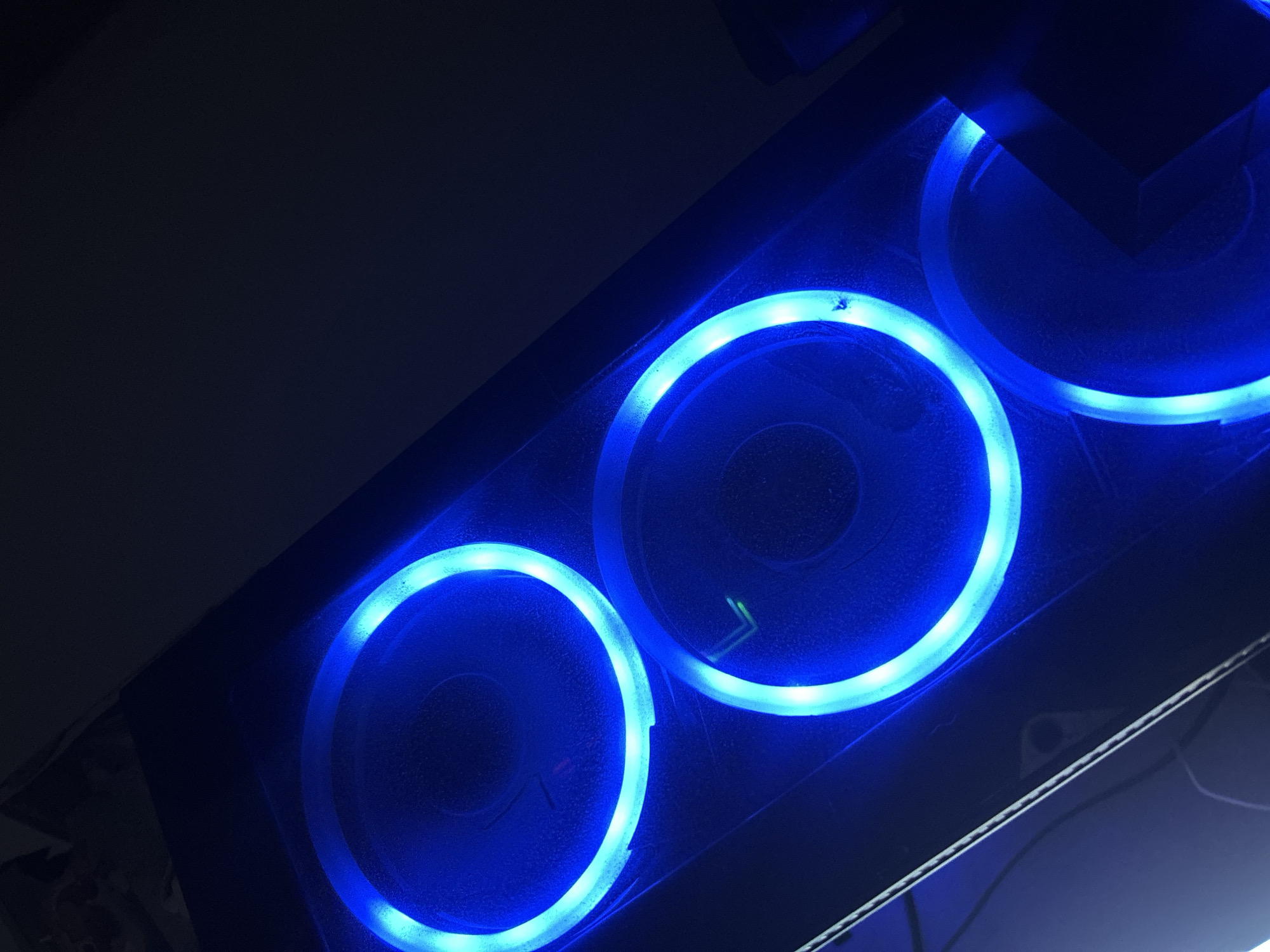 A close-up, angled view of a computer case with three blue LED-lit fans. The fans are circular, with the LED rings emitting a bright blue light. The computer case appears to be dark-colored. The environment is dark. There are no people visible. The location is unknown and cannot be determined from the image.