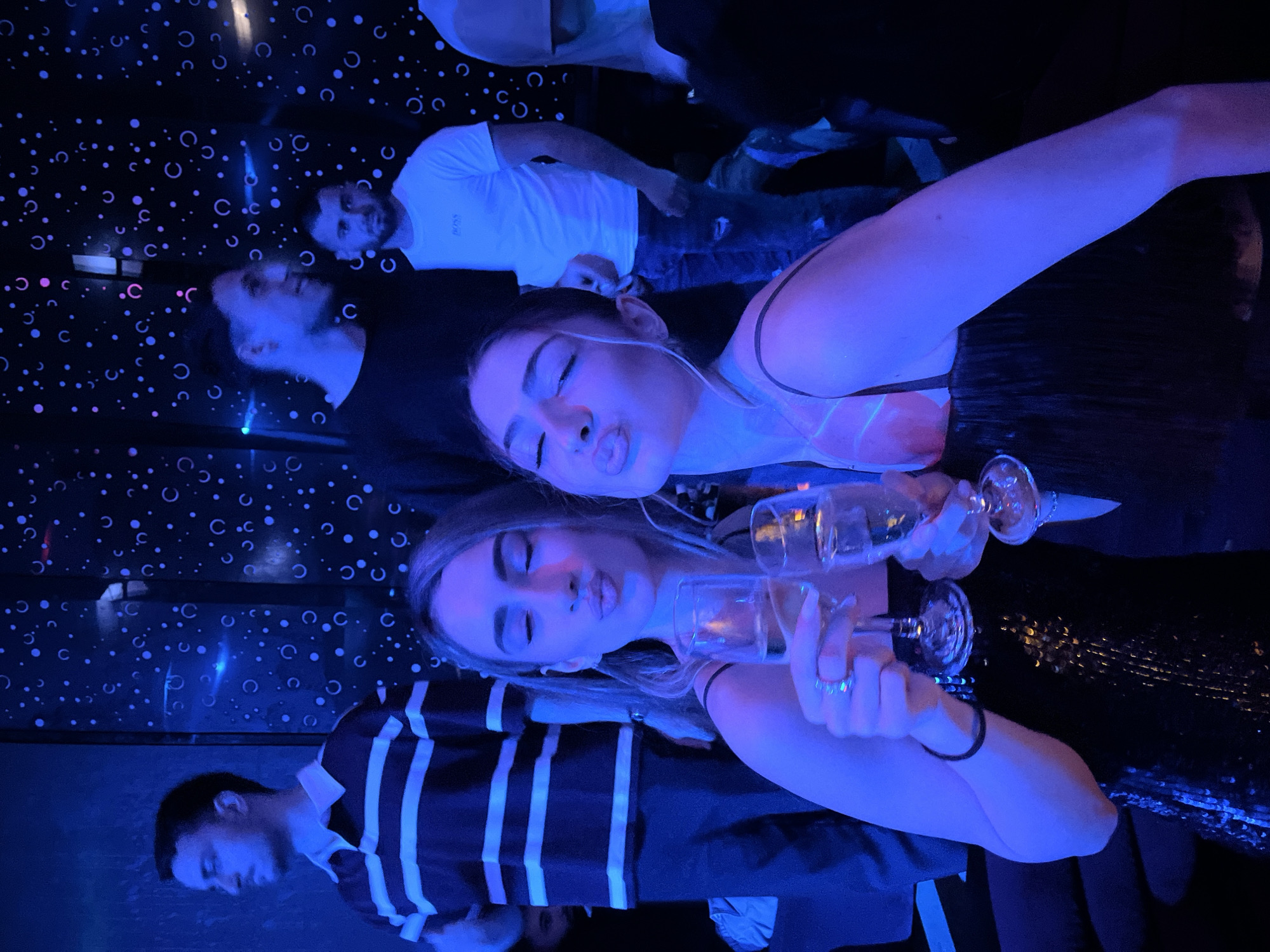 The image depicts a group of people posing with champagne glasses in what appears to be a nightlife setting. Two women in the foreground hold the glasses, making a kissing face at the camera. They wear what looks like party attire. One wears a dress with sequins or a similar shimmering texture. Behind them and to the left, a man is wearing a black and white striped shirt. Further back, near the wall, two more men are visible. One is wearing a white shirt, the other is wearing a black shirt. The location seems to be indoors, with a wall or backdrop decorated with circular patterns. A blue light filters across the whole scene. There is not enough information to provide the location in Burgas, Bulgaria.