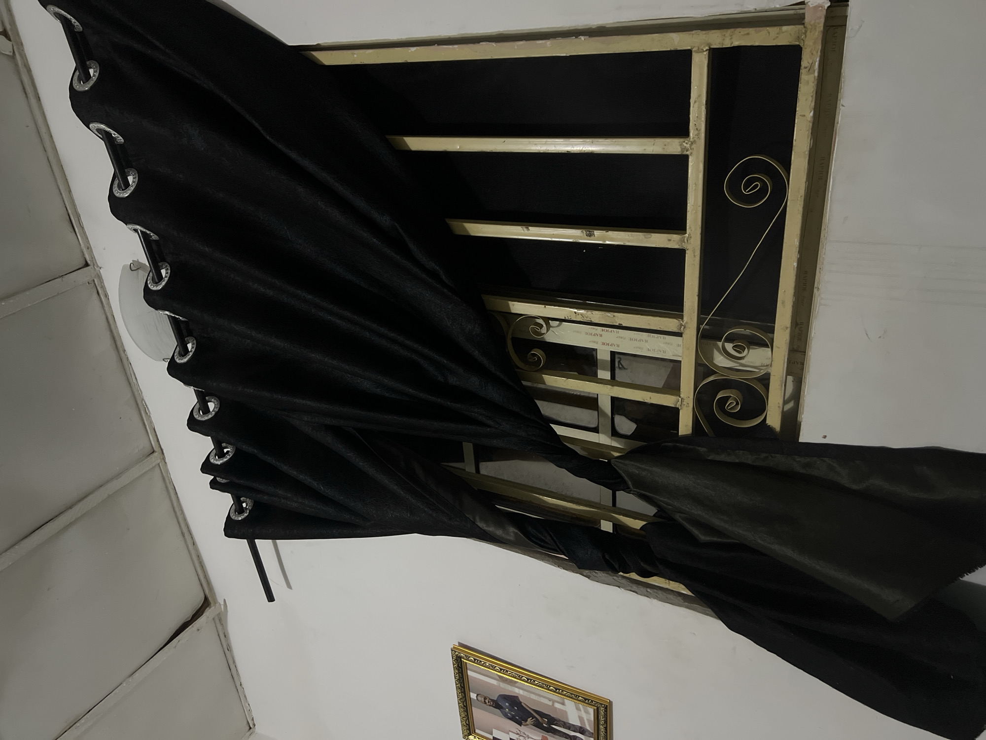 The image depicts an indoor scene. A black curtain with grommets is drawn across a window with a metal grate. The grate is a light gold color and features decorative scrollwork. Behind the grate, a black mesh screen is visible. To the left of the window, a framed portrait of a man in a dark suit is hanging on a white wall. The ceiling, visible at the top of the image, appears to be made of white panels. The overall impression is of a room in a residential setting in Howayi, Nigeria.
