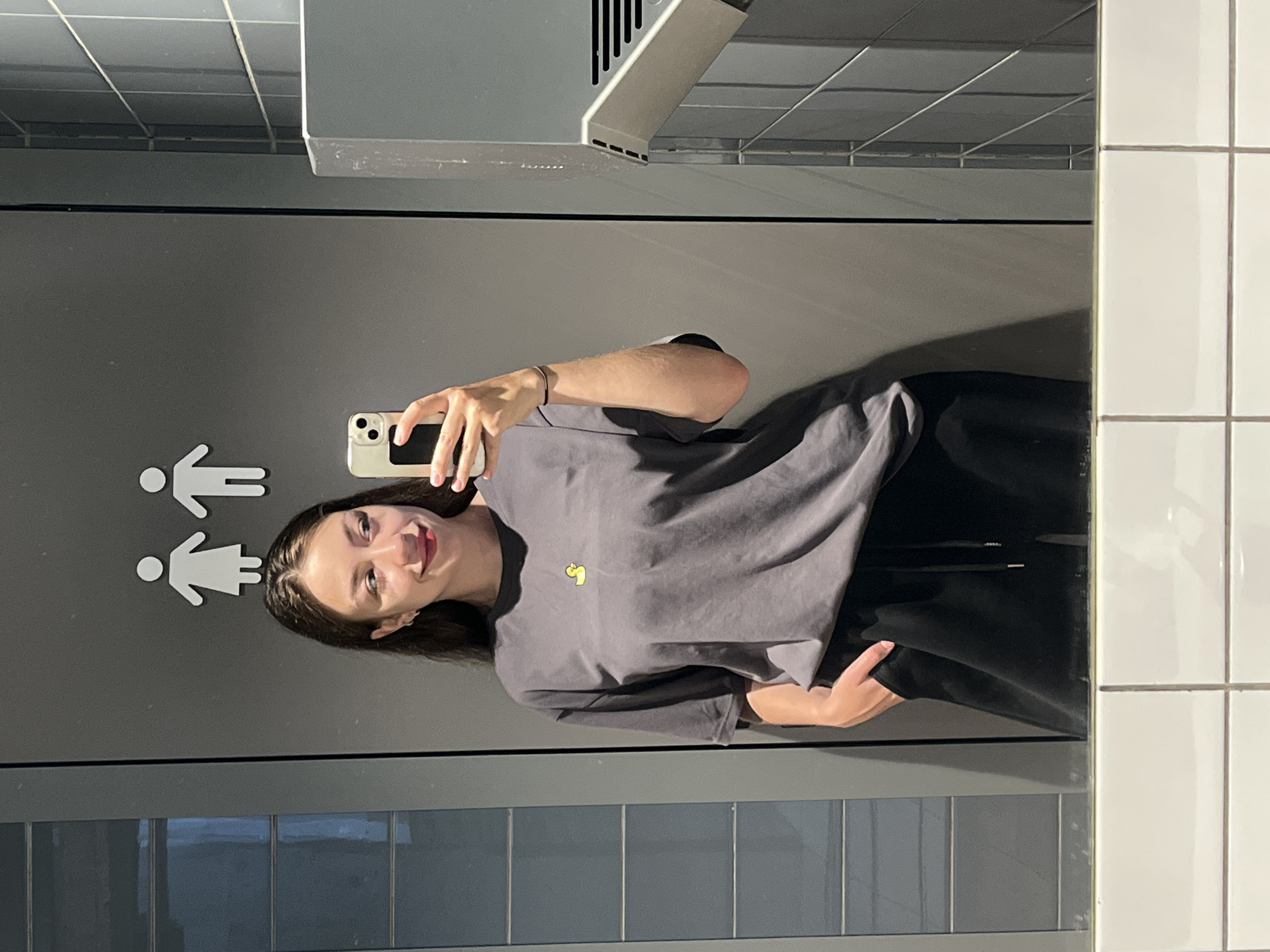 A young woman takes a mirror selfie in a public restroom. She is wearing a dark grey t-shirt with a small yellow duck logo and black sweatpants. Her right hand is in her pocket. She holds a white smartphone in her left hand, angled to capture her reflection. The background consists of a grey tiled wall, a restroom sign with male and female figures, and a hand dryer. The floor is tiled with white squares. The image does not provide any information about the city of Chaikovsky, Russia, or any other specific location.