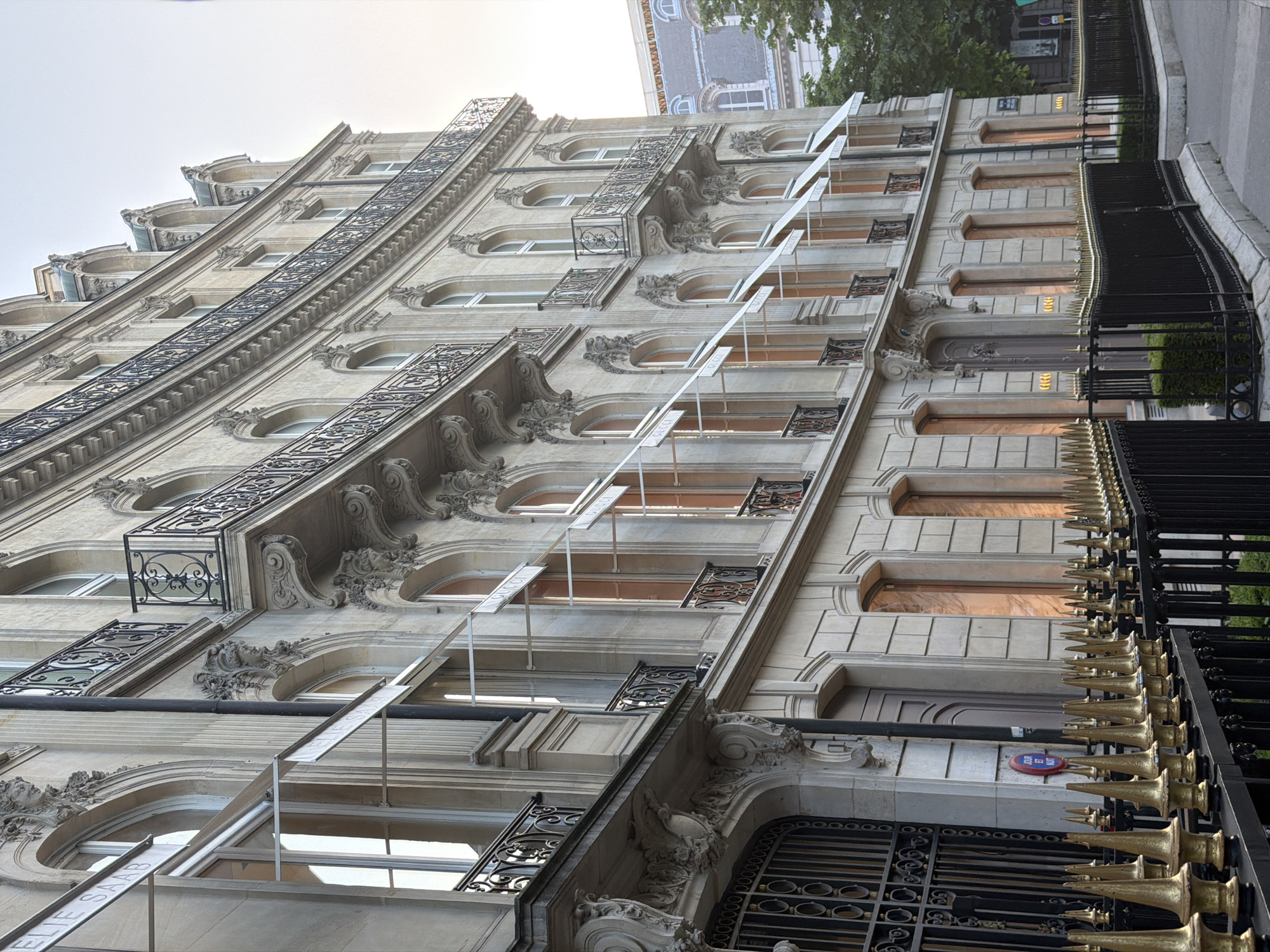The image shows a low-angle view of a multi-story building in Paris, France. The architecture is ornate, with decorative molding, arched windows, and wrought-iron balconies. Vertical signs are attached to the building at each window, indicating designer brands. A wrought-iron fence with gilded spear-shaped finials runs along the sidewalk in front of the building. The street and a glimpse of greenery are visible in the background.