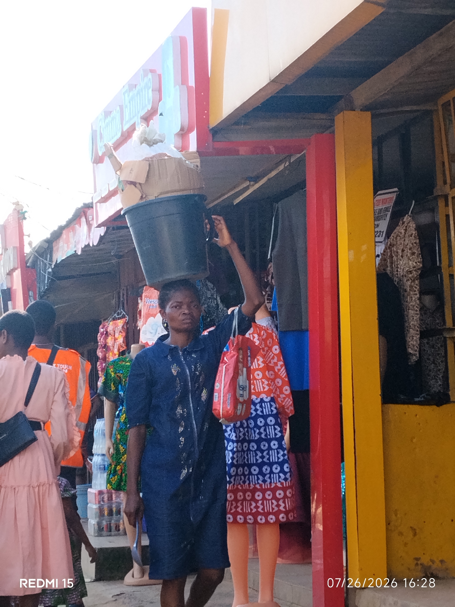 In Umuode, Nigeria, a woman with dark skin and dark hair is pictured carrying a large black bucket filled with cardboard boxes on her head. She is wearing a dark blue denim jacket with intricate patterns and a dark blue skirt. Her left arm is raised, supporting the bucket. In her right hand, she holds a silver spoon or utensil. In front of her, a mannequin wearing a red and white patterned dress is visible. Behind the woman, another woman in a light pink dress with ruffles on the sleeves is seen carrying a black purse. To the left of the main subject, a person wearing an orange reflective vest can be seen. The scene appears to be a market or roadside shop with stalls displaying clothing. The background features colorful building structures, including red and yellow pillars, and signs with text, one of which reads "COMIC EMPIRE". The image also has text overlays that say "REDMI 15" and "07/26/2026 16:28".