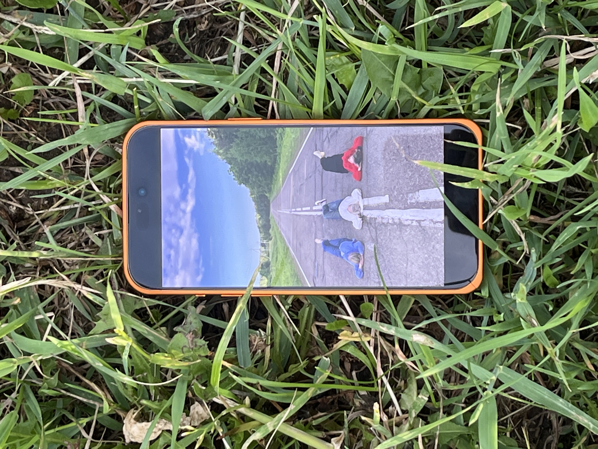 An overhead shot depicts an orange-cased smartphone resting horizontally on a bed of green and brown grass. The grass blades are varied in length and texture, suggesting a natural outdoor environment. The phone's screen is illuminated, displaying a vibrant image rotated 90 degrees counter-clockwise relative to the phone's orientation.
The image on the phone screen features an asphalt road stretching into the distance under a bright blue sky with scattered white clouds. Green trees and foliage line both sides of the road. Three individuals are lying on their backs on the road's surface, arranged along the white dashed line that divides the road. One person wears a red top and black pants, positioned towards the top right of the screen image. Another, in light-colored clothing, lies closer to the center-left. The third individual, dressed in blue, is situated towards the bottom left. All three appear to be looking upwards, seemingly at the sky within the displayed image.
The main image is taken outdoors during what appears to be daytime, given the clear illumination. There is no visible text in the image. The scene is set in Bol'shoi Bukor, Russia.