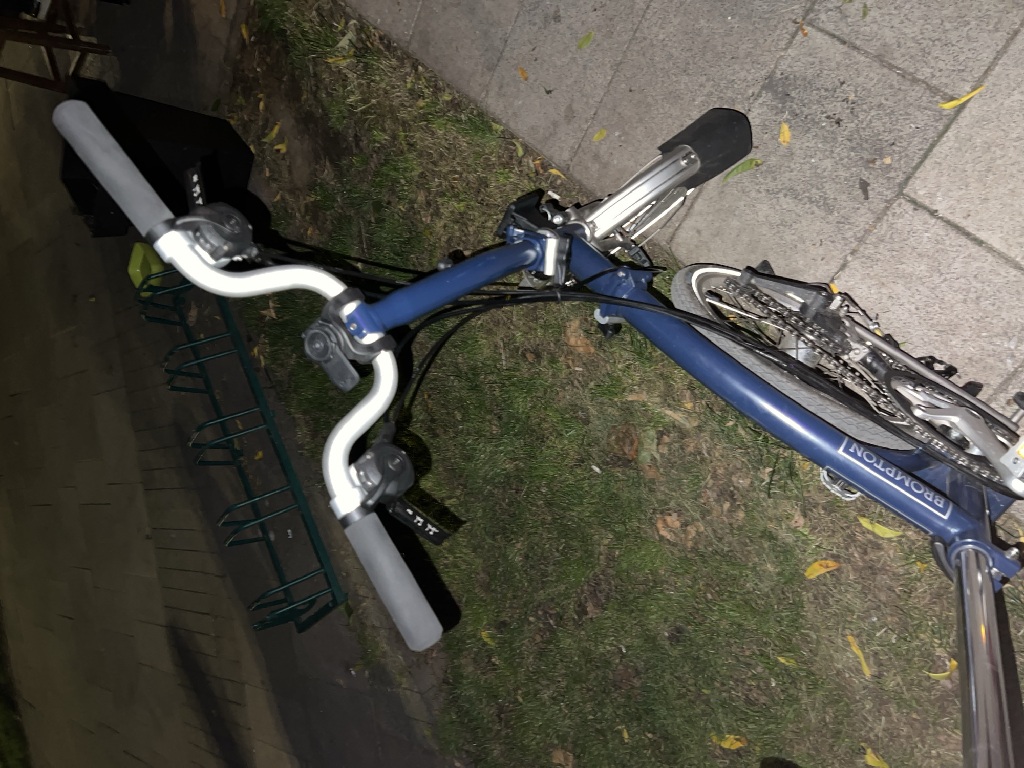 A blue Brompton folding bicycle is parked on a paved sidewalk next to a patch of grass. The bicycle is viewed from a high angle, looking down on the handlebars and front of the bike. The handlebars are silver with grey grips. There are various components visible on the handlebars, including what appear to be brake levers and gear shifters. A black basket is attached behind the handlebars. The bicycle frame is a deep blue color and has the "Brompton" logo printed on it in white text. The bicycle's front wheel and part of the chain and gears are visible. In the background, there is a green bicycle rack and a tiled walkway. Fallen yellow leaves are scattered on the grass and sidewalk. The scene appears to be outdoors in an urban setting. There are no people present in the image. The lighting suggests it may be dusk or night, with artificial light illuminating the scene. The location is identified as Sofia, Bulgaria.