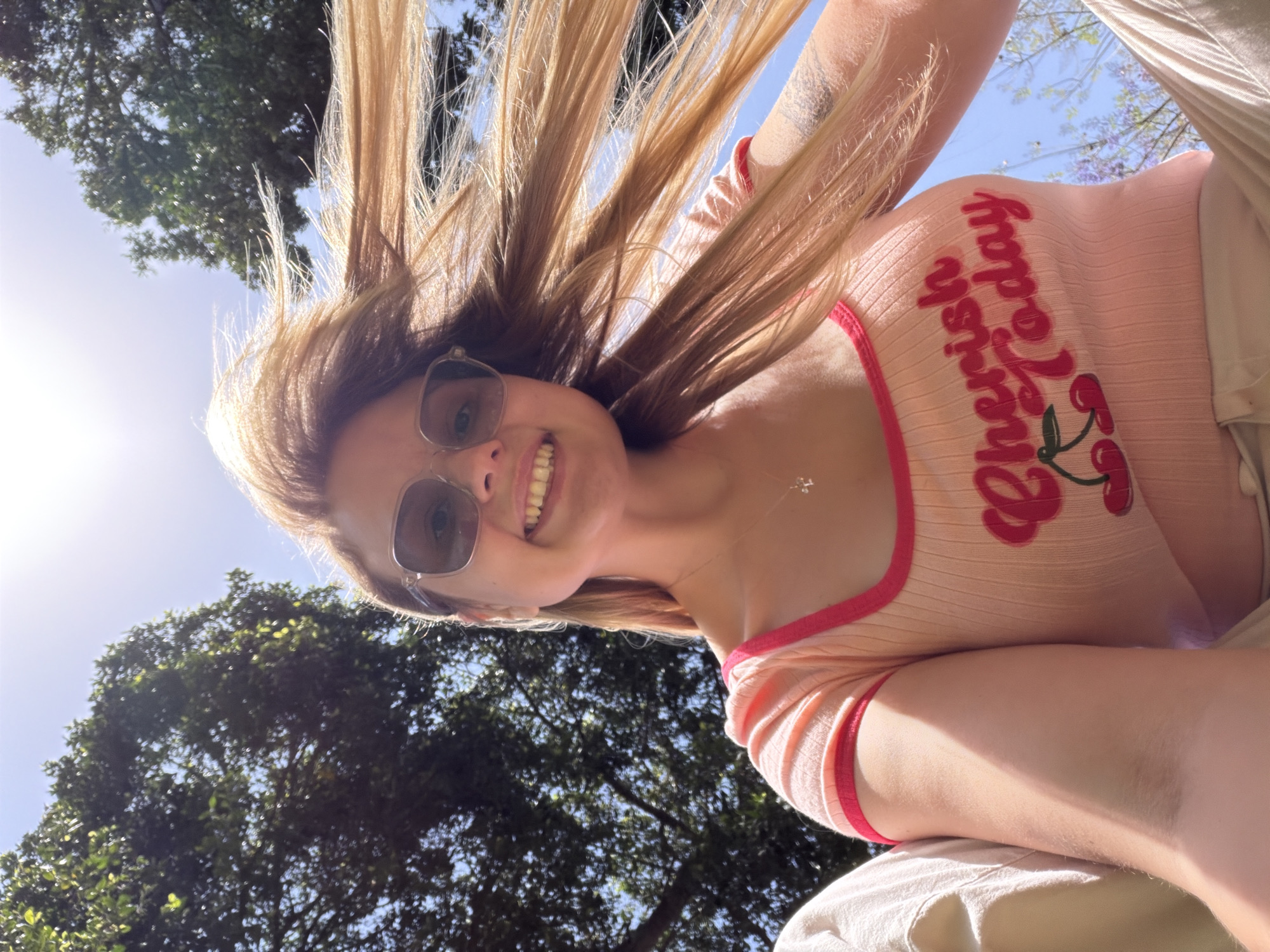 A woman is photographed from a low angle, seemingly lying on her back outdoors. She wears sunglasses and a pink top with red trim and the words "Cherish Today" printed on it with an image of cherries. Her long blonde hair is swept back, possibly by the wind or her position. The sky is visible behind her with trees and the sun. No information indicates this is specifically in Tel-Aviv.