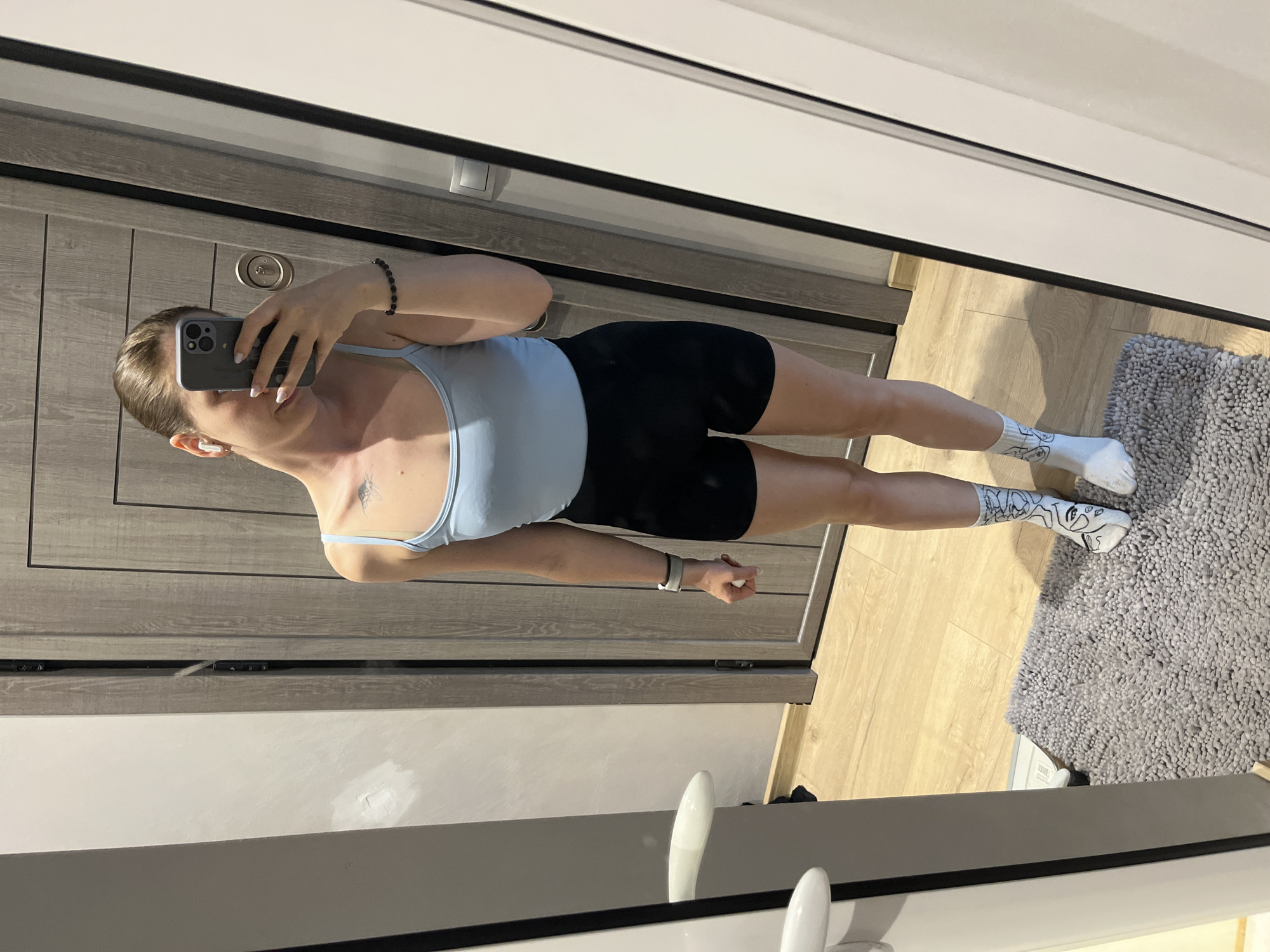 A young woman stands in front of a mirror, taking a selfie in what appears to be a domestic setting, possibly a bathroom or dressing area. She is wearing a light blue sleeveless top and black shorts, with white socks that have a black line drawing design on them. She has a bracelet on her left wrist and what looks like a smartwatch. She is holding a smartphone in her right hand, which is angled to capture her reflection. In the background, a wooden door with vertical panels is visible, along with a grey rug on a wooden floor. There is also a silver-colored scale visible on the floor to the left of the rug. The scene does not provide any specific indicators of its location within Pazardzhik, Bulgaria.