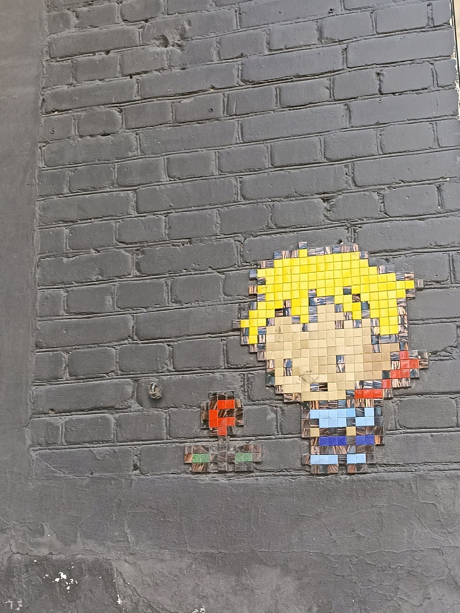 The image shows a pixel art mosaic of a cartoon character affixed to a dark gray brick wall. The mosaic depicts a boy with spiky yellow hair, light brown skin, a red bandana, and a blue and white striped shirt. To the left of the boy is a smaller, abstract mosaic with red and green pixelated elements. The brick wall has visible mortar lines and appears to be aged. The bottom portion of the image shows a rougher, darker textured surface that may be part of the same wall or a different structure.
This mosaic is located in Novosibirsk, Russia, specifically on the wall of the Novosibirsk State Museum of Local Lore.