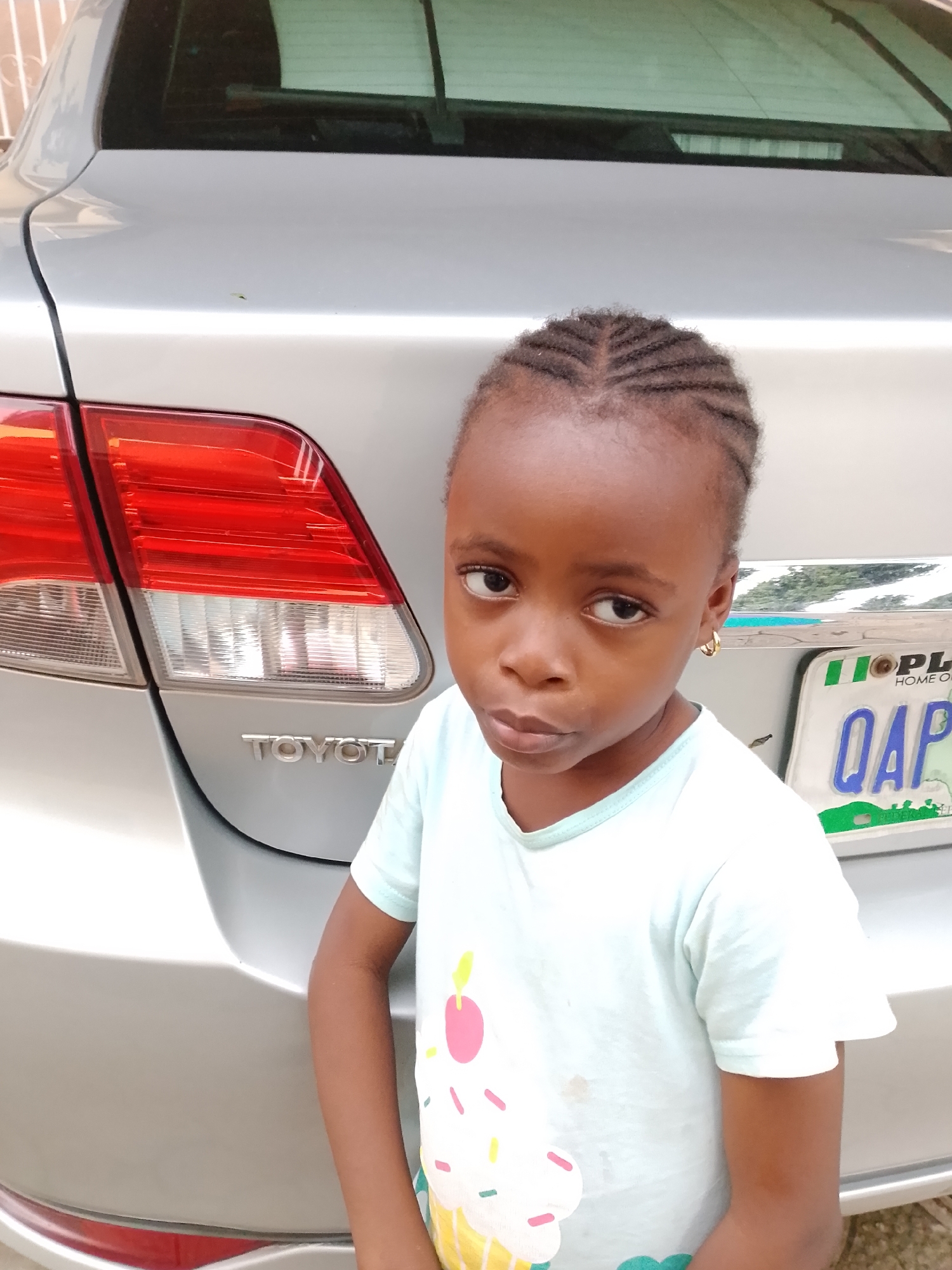 A young girl stands in front of a silver Toyota sedan in an outdoor setting, likely a residential area given the presence of a fence in the background. The girl is facing the camera, with her left side slightly closer. She has dark skin, braided hair, and is wearing a light blue t-shirt with a cupcake graphic. She has a serious expression on her face. Behind her, the rear of the car is visible, including the taillights and a license plate with the letters "QAP". The license plate appears to be from Nigeria, indicated by the flag colors and "HOME OF PEACE" inscription. The lighting suggests it is daytime, possibly with overcast conditions.