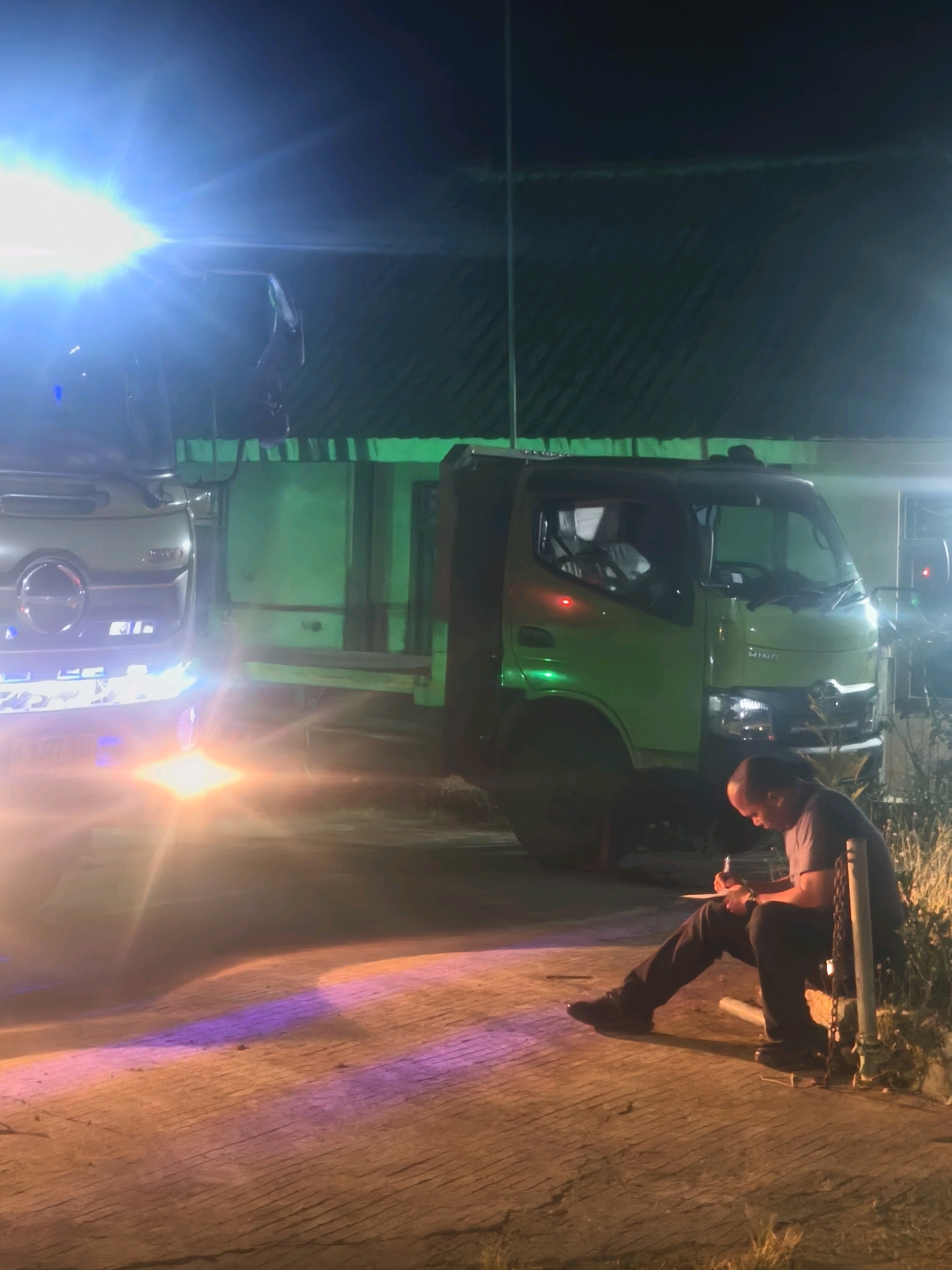 The image depicts a nighttime scene at a location in the city of Kroya, Indonesia. A man is seated on the ground, leaning against a metal pole, and appears to be writing on a pad of paper. He is dressed in dark clothing. In the background, two trucks are parked. One truck, prominently positioned in the center, is green and has its headlights on. To its left, another truck, lighter in color, also has its headlights illuminated, casting bright light and lens flares across the scene. The ground is paved and appears wet in places, reflecting the colorful lights from the trucks. The overall ambiance is illuminated by artificial lighting, creating a somewhat dramatic and potentially industrial or logistical setting.