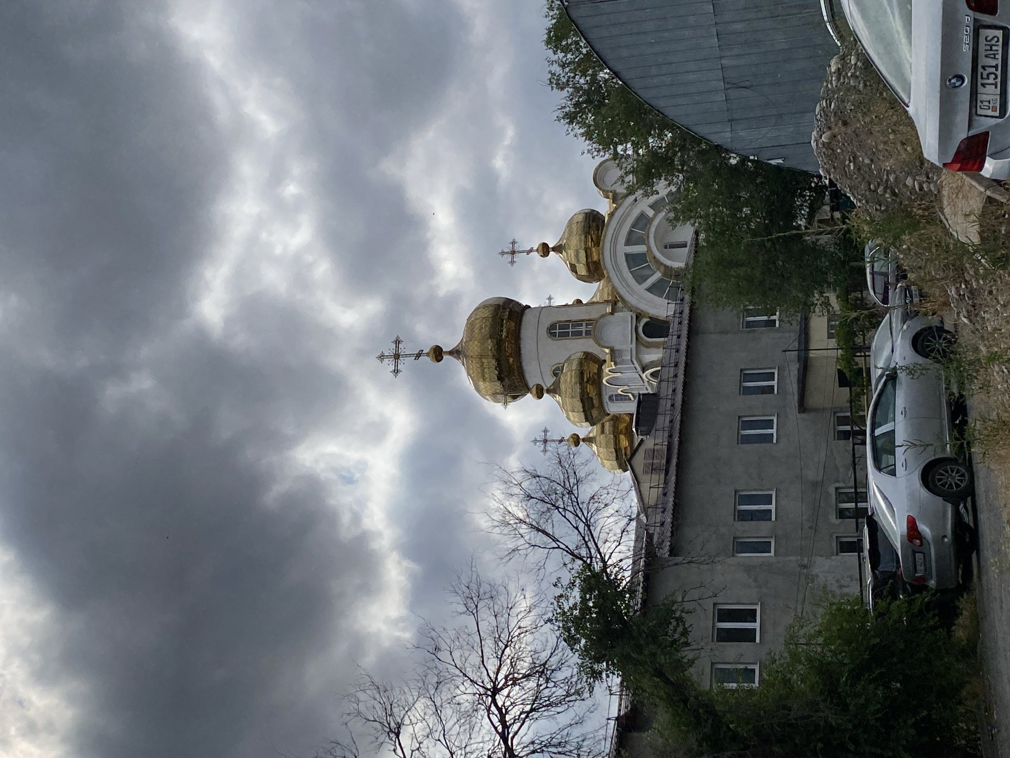 The image shows a street view of a building with golden domes and crosses, likely a church or cathedral, in Bishkek, Kyrgyzstan. The sky is overcast with dark clouds. In the foreground, there is a gray building with several windows, and parked cars. There are trees with bare branches on the left side of the image. The overall scene suggests an urban setting under a cloudy sky.