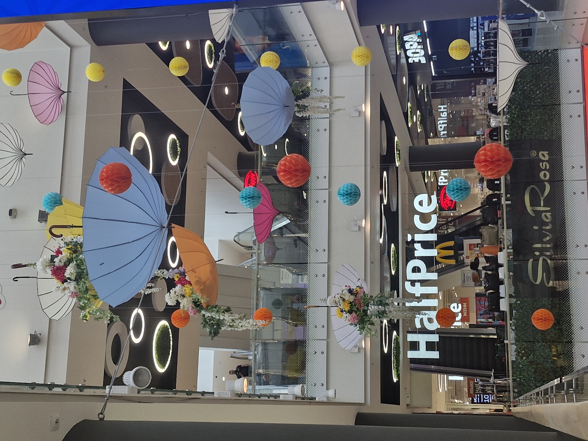 This image captures the interior of a brightly lit, multi-story shopping mall, likely in Sofia, Bulgaria, given the presence of Cyrillic text. The perspective is from an upper level, looking down into a large atrium or open space.
The main subjects are the extensive and colorful decorations hanging from the ceiling. Numerous umbrellas in various colors such as pink, light blue, yellow, orange, and white are suspended upside down. Many of these umbrellas are adorned with artificial flowers in white, pink, and yellow. Interspersed among the umbrellas are spherical honeycomb paper decorations in vibrant hues of yellow, orange, red, and turquoise. These decorations create a cheerful and whimsical atmosphere, suggesting a festive season or a special event.
In the background, multiple retail store facades are visible on different levels. Escalators can be seen connecting the floors. Circular and oval-shaped light fixtures, some incorporating greenery, decorate the walls and pillars, contributing to the modern aesthetic of the mall. Reflections on glass barriers and shop windows are also noticeable.
Visible text includes the prominent store names "HalfPrice" and "Silvia Rosa." The yellow "M" logo for McDonald's is also discernible. Cyrillic text, such as "ЖЕНИ" (Women) and "МЪЖКЕ" (Men's), likely indicating departments or restrooms, can be seen on lower levels. Another partially visible sign reads "10 ЗОН" (10 Zones).
A few indistinct figures of people, likely shoppers or mall staff, are visible in the distance on the lower levels, but no specific activity can be discerned beyond general mall operations. The lighting suggests it is either daytime with ample natural light from above or a well-lit indoor environment.