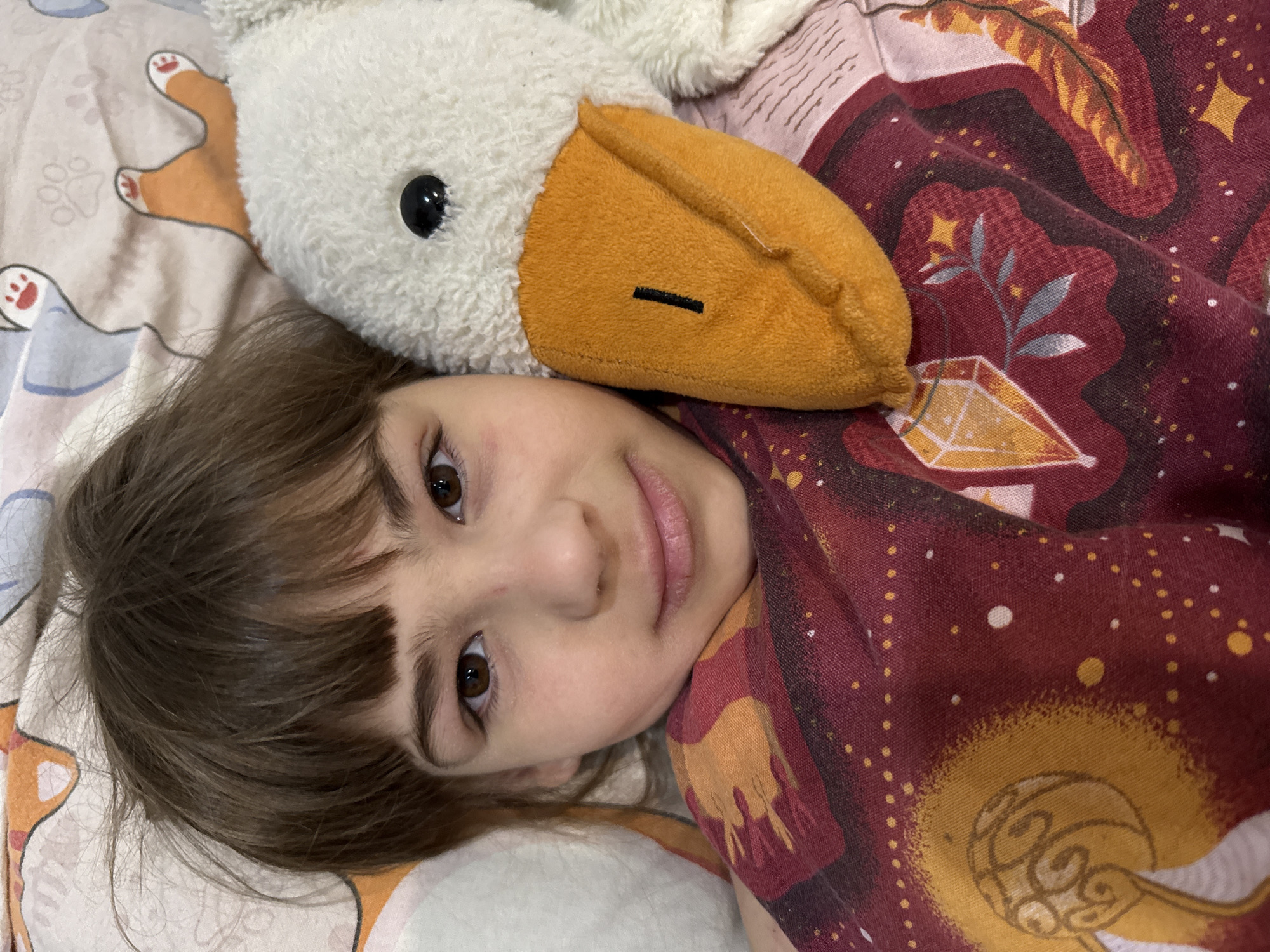 A young child with brown eyes and bangs lies on a bed in what appears to be a home environment. A plush toy resembling a bird, possibly a goose, is positioned near the child's head. The bed has patterned sheets with drawings of paws and bones, while the child is covered in a blanket or wearing a shirt with a dark red and gold mystical pattern of stars, crystals, and botanical elements. The image provides no specific details to confirm the location is in Шебунино, Russia.